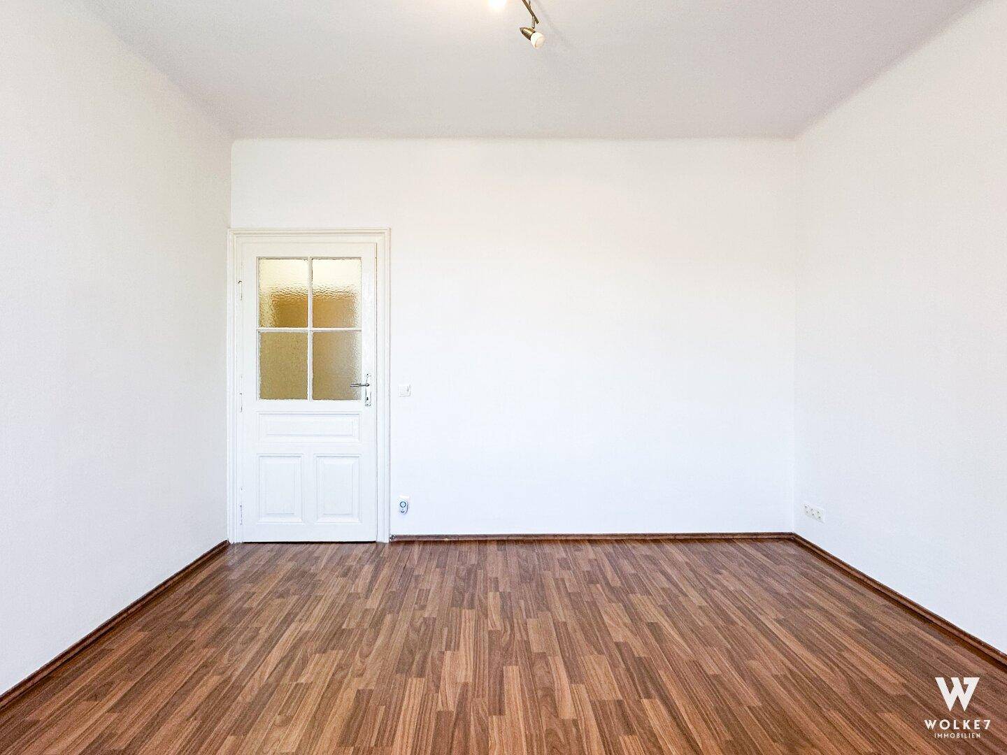 Leerer Raum mit weißen Wänden und hellem Laminatboden, bereit für die individuelle Gestaltung.