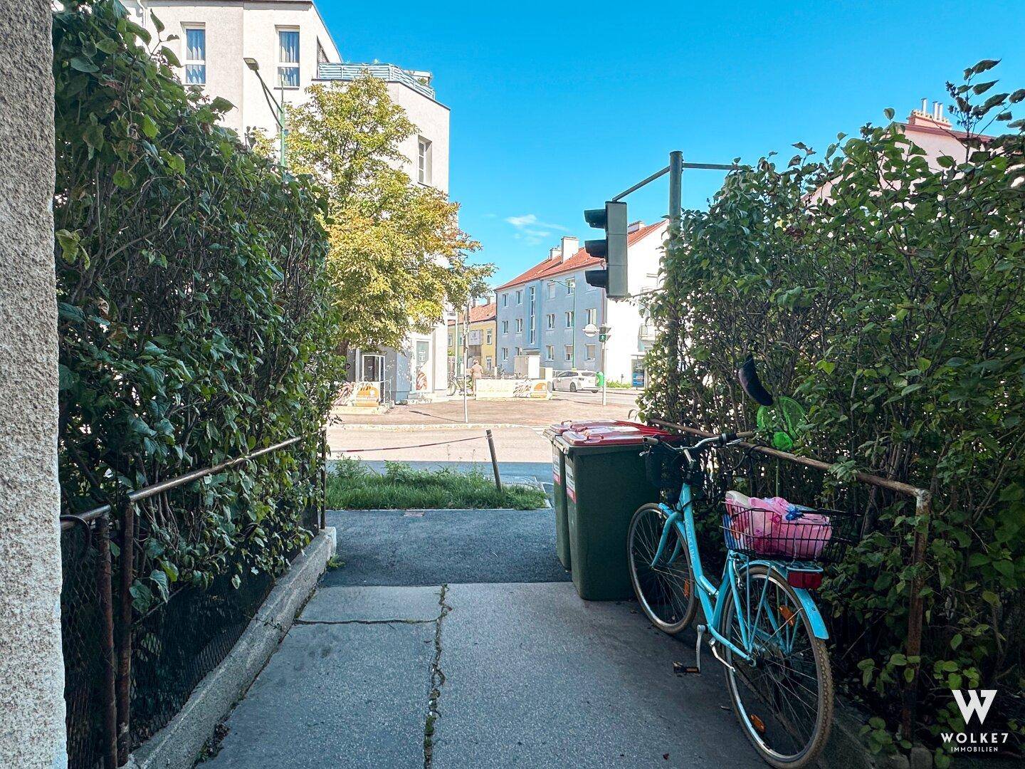 Blick von einem schmalen Weg zwischen Hecken auf eine Wohnstraße mit Gebäuden und Fahrrädern.