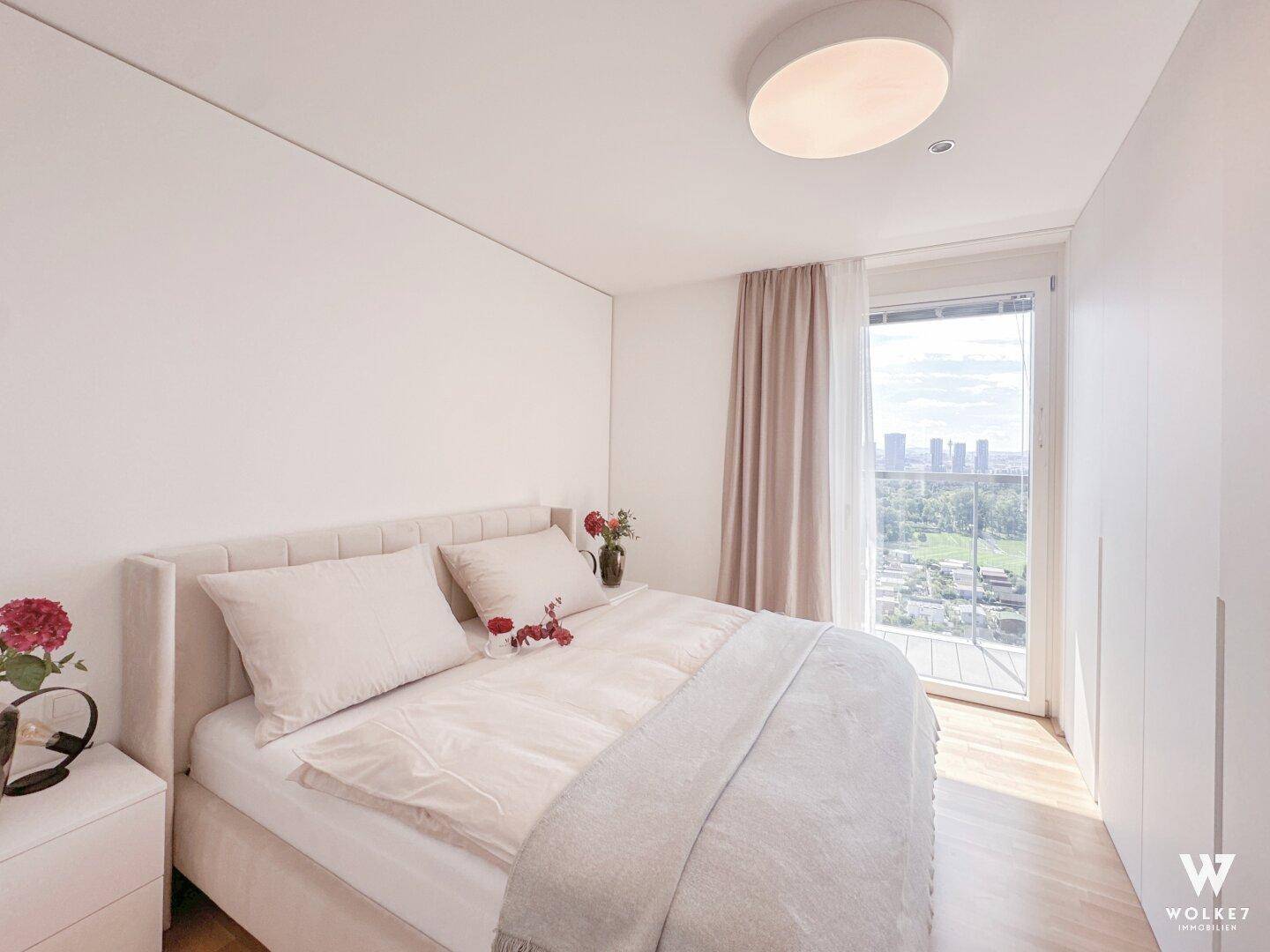 Helles Schlafzimmer mit Doppelbett, schlichtem Design und großem Fenster mit Stadtblick.