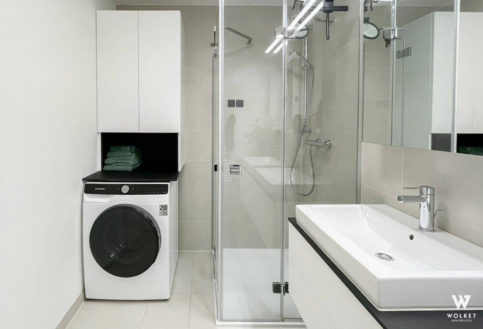 Helles Badezimmer mit Dusche, Waschbecken und integrierter Waschmaschine.