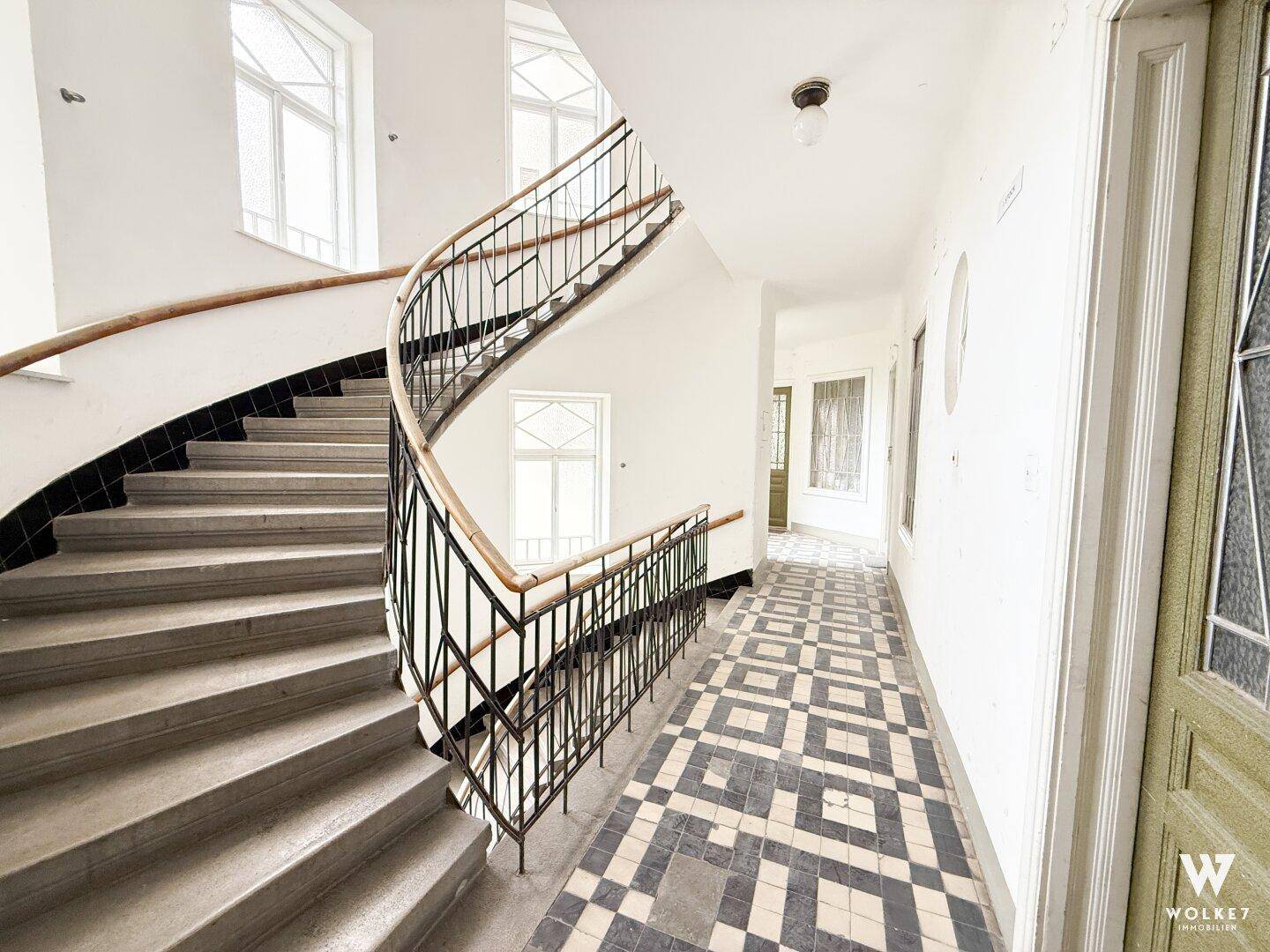 Historisches Treppenhaus mit geschwungener Treppe, schmiedeeisernem Geländer und gemusterten Fliesen im Flurbereich.