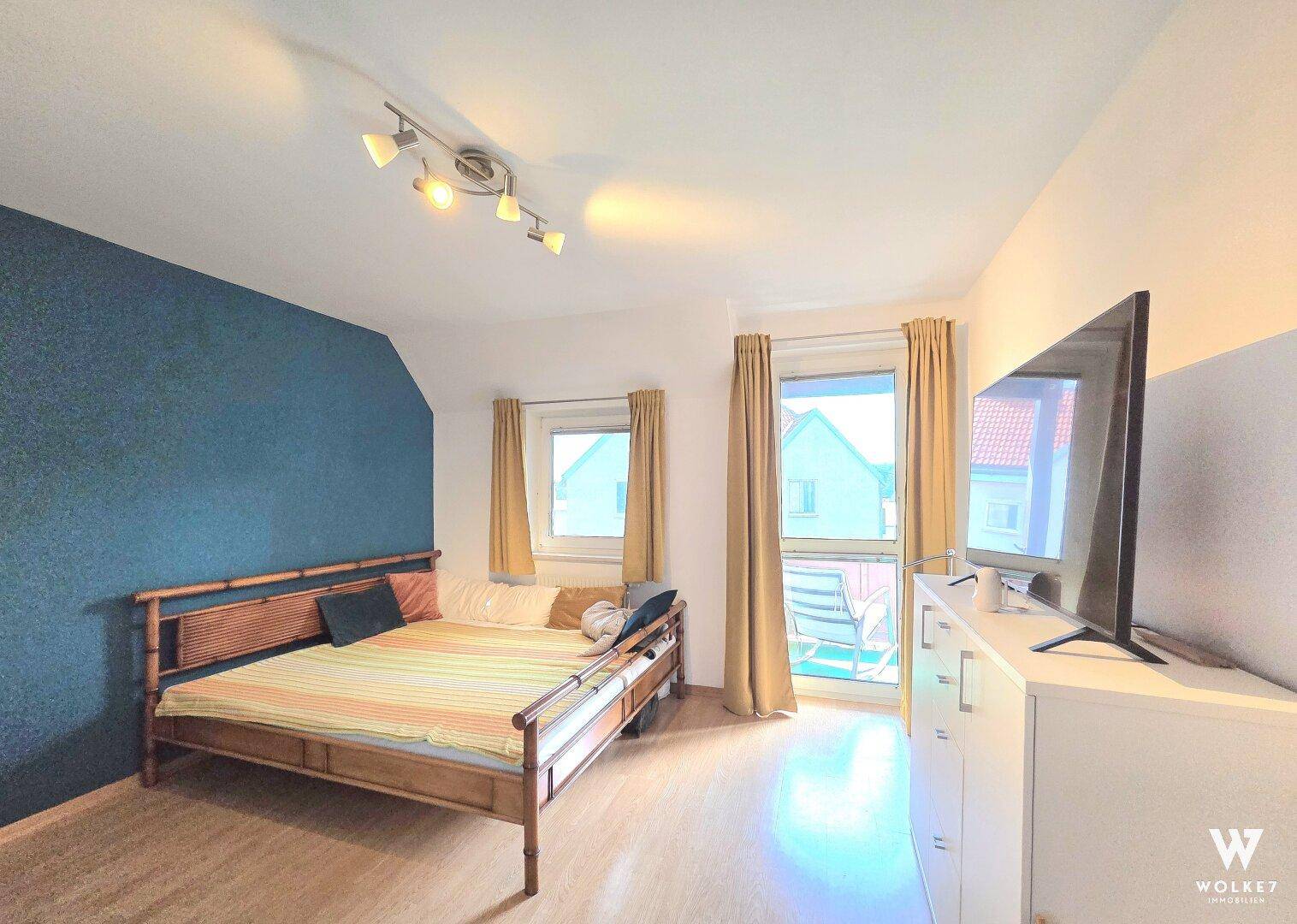 Schlafzimmer mit Holzbett, blauer Akzentwand und Zugang zum Balkon.