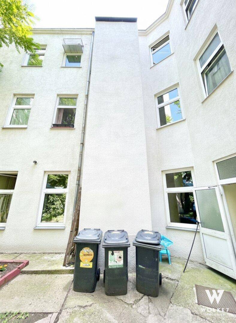 Hinterhofbereich eines Mehrfamilienhauses mit weißer Fassade, Fenstern und Mülltonnen.