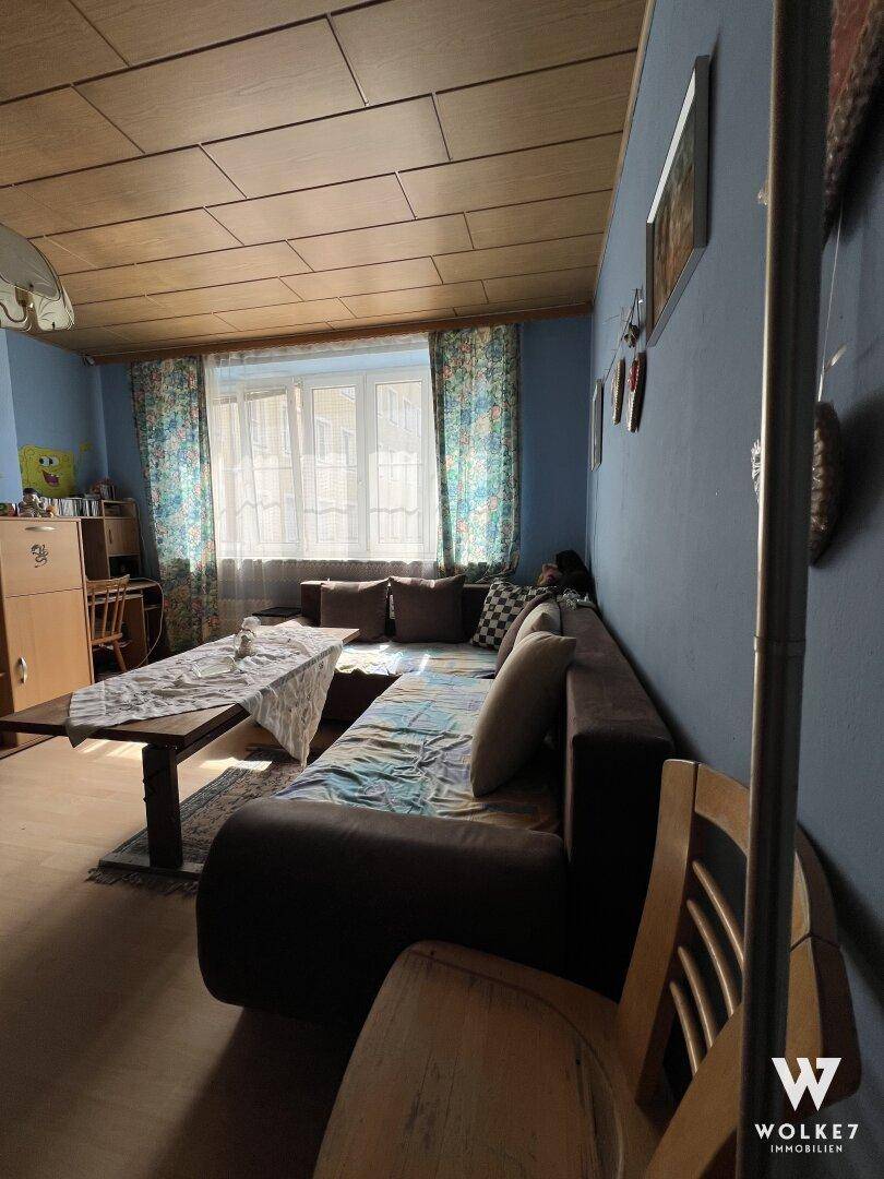 Gemütliches Wohnzimmer mit Sofa, Couchtisch, Holzdeckenverkleidung und bunten Vorhängen am Fenster.