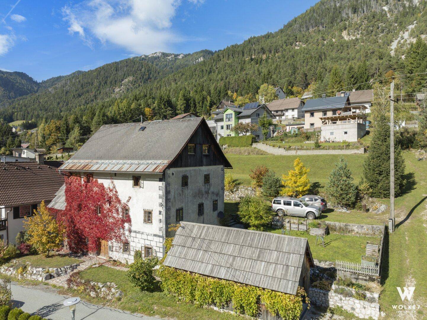 Historisches Gebäude mit rotem Efeu bewachsen, umgeben von grüner Landschaft und Bergen.