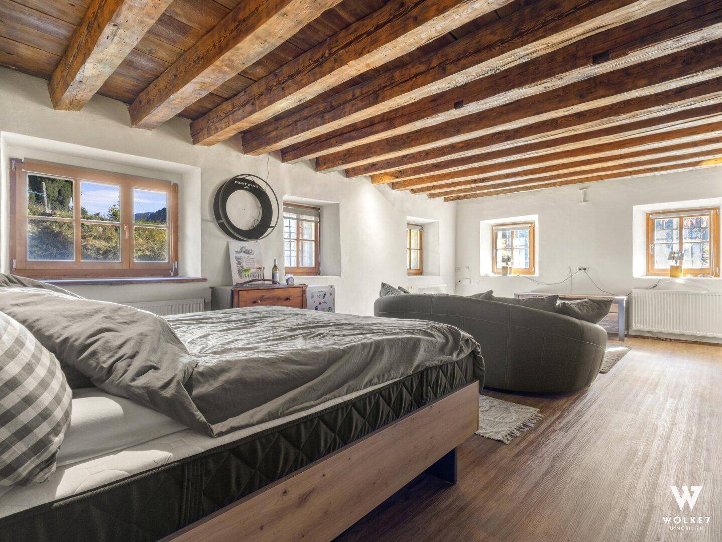 Helles Schlafzimmer mit Holzbalkendecke, großem Bett und Blick auf die grüne Umgebung.