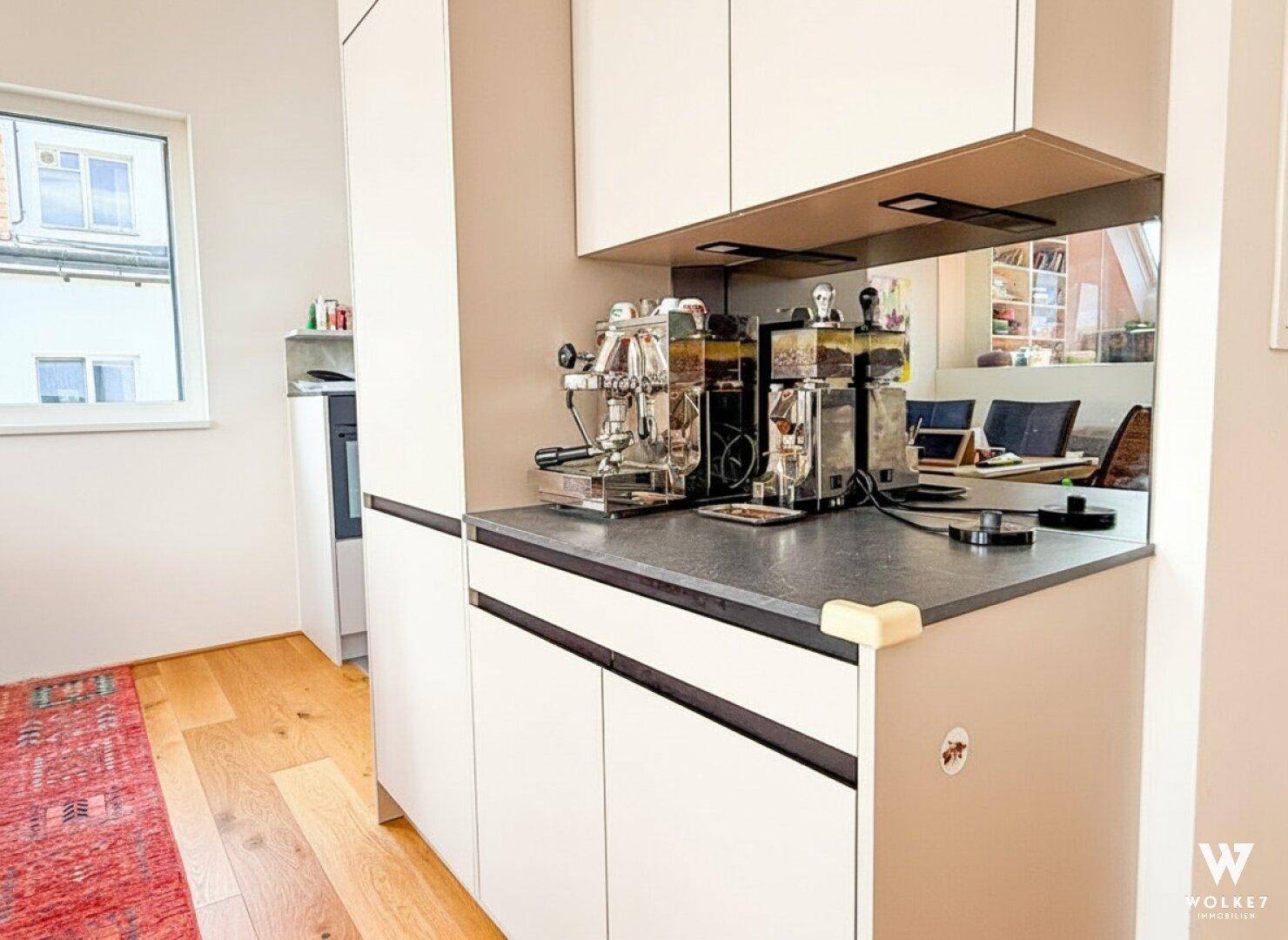 Küchenzeile mit integrierter Kaffeemaschine, weißen Schränken und einem Fenster, das den Blick nach draußen freigibt.