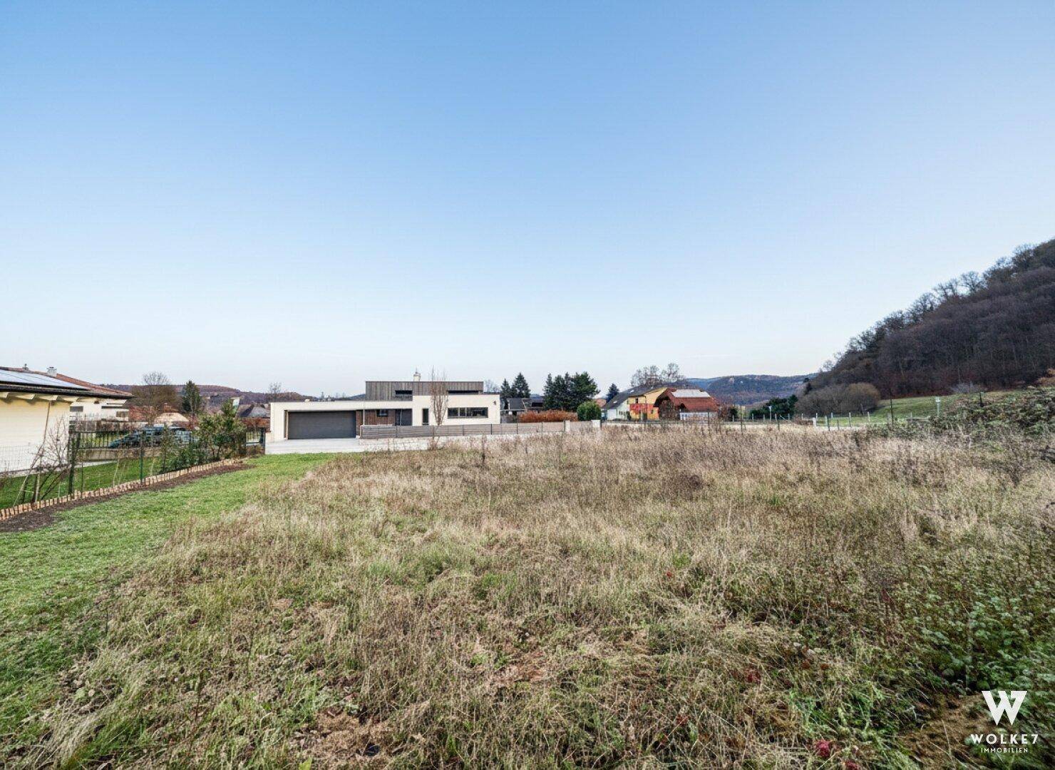 Unbebautes Grundstück mit Blick auf moderne Wohnhäuser und bewaldete Hügel in der Ferne.