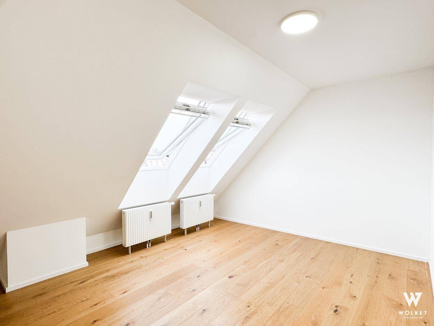 Leeres Zimmer im Dachgeschoss mit Holzboden, Heizkörpern und viel Tageslicht durch Dachfenster.