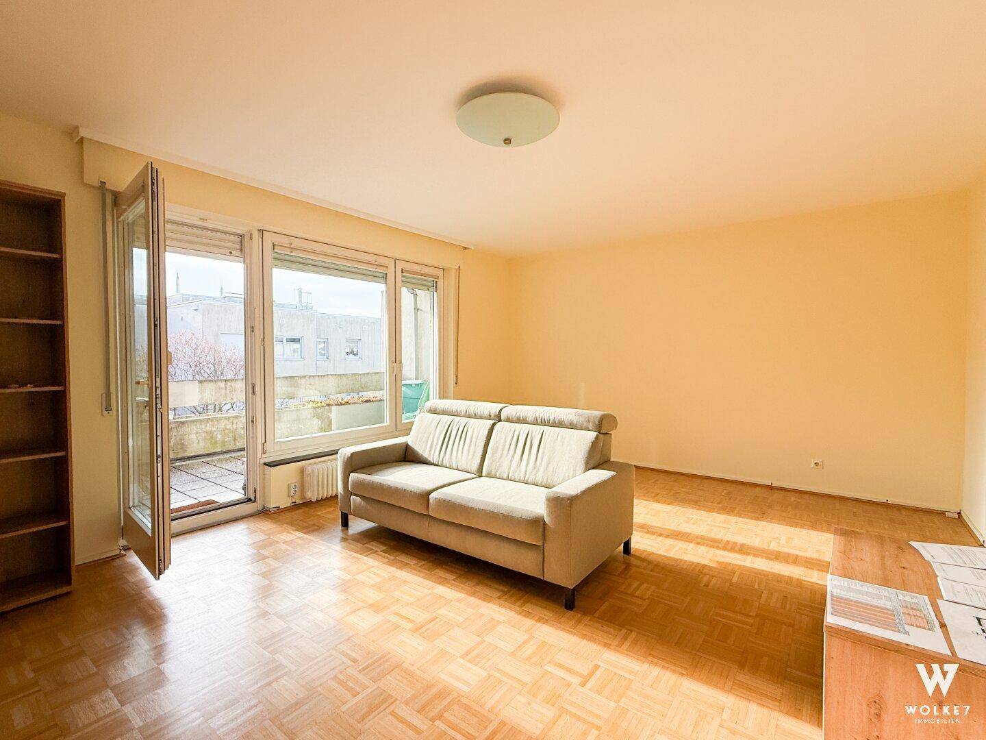 Helles Wohnzimmer mit Parkettboden, beiger Couch und Zugang zu einem Balkon oder einer Terrasse.