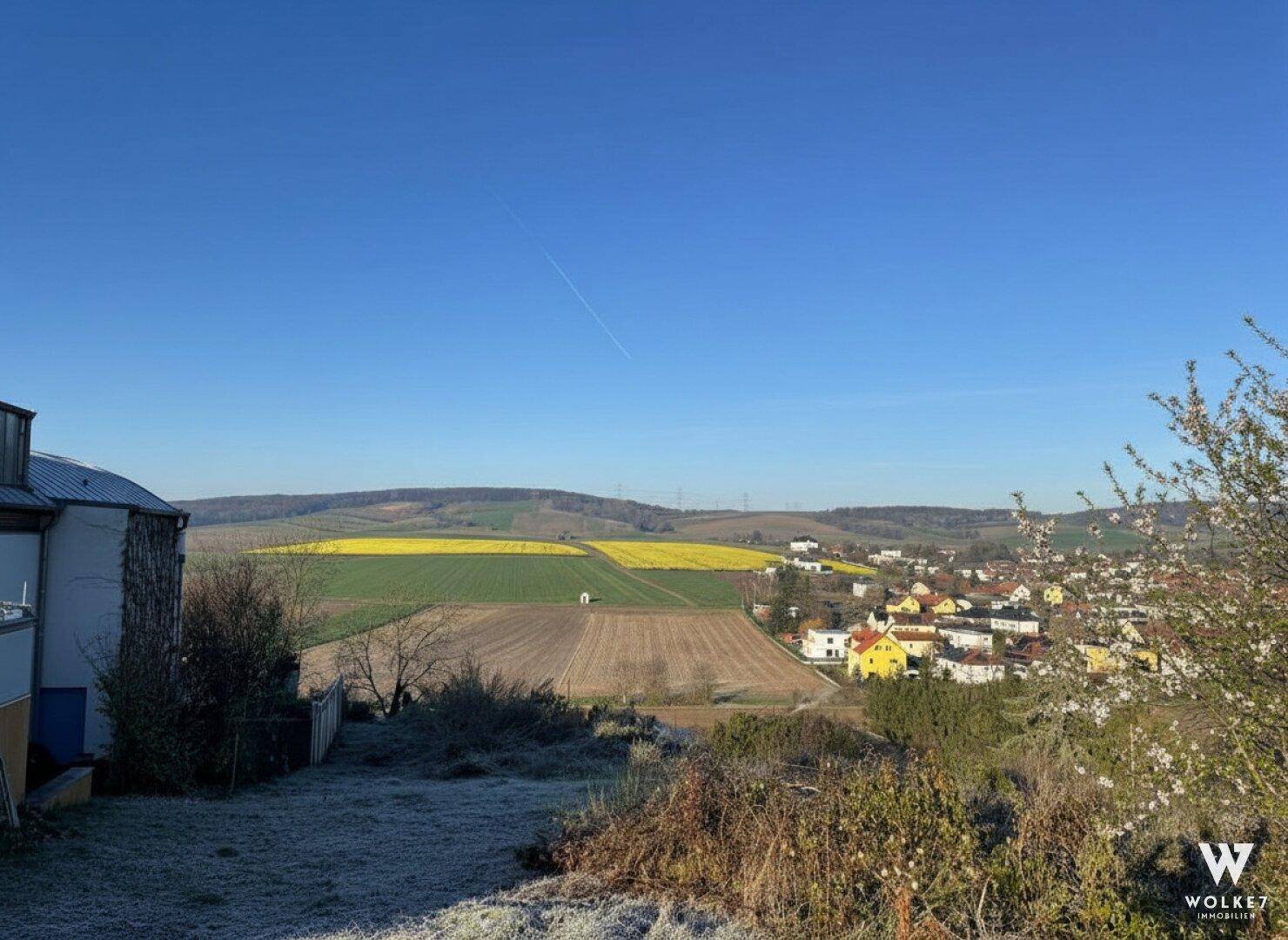 Weitläufiger Ausblick auf eine ländliche Umgebung mit Feldern, einem Dorf und einem Gebäude im Vordergrund.