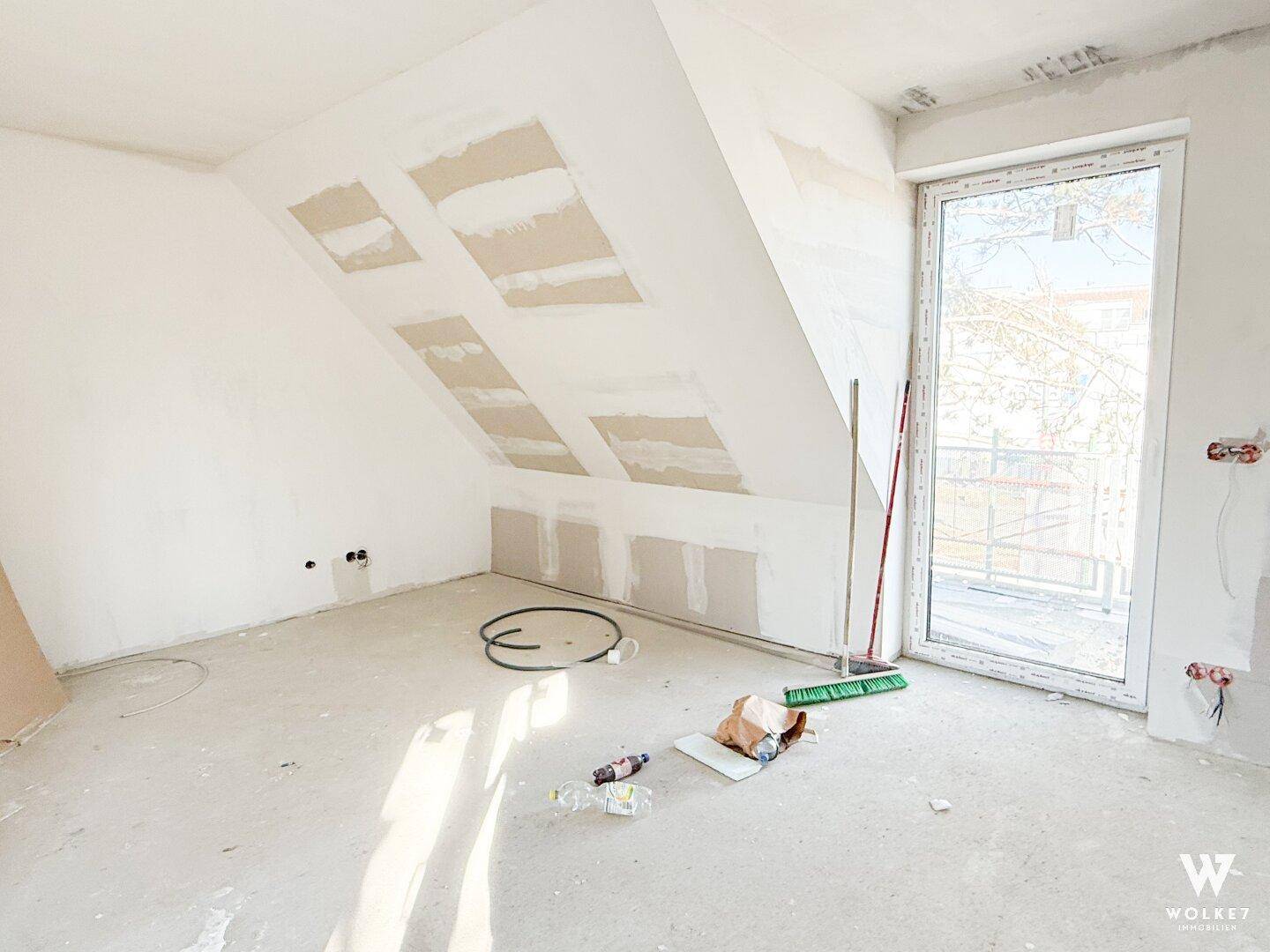 Dachgeschosszimmer im Rohbau mit schräger Decke und Zugang zu einem Balkon.