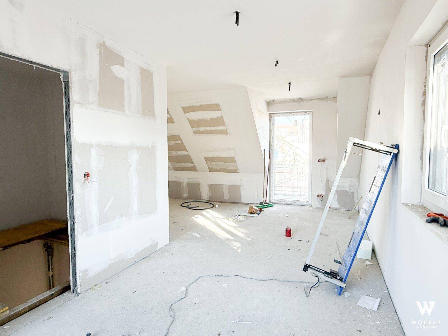 Dachgeschossraum im Rohbau mit schrägen Wänden und Fenster, bereit für den Ausbau.