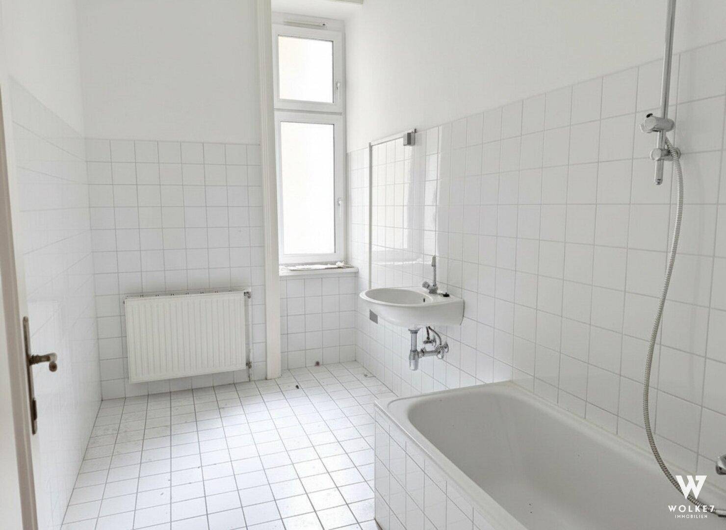 Helles Badezimmer mit weißen Fliesen an Boden und Wänden, einer Badewanne, Waschbecken und einem Fenster.
