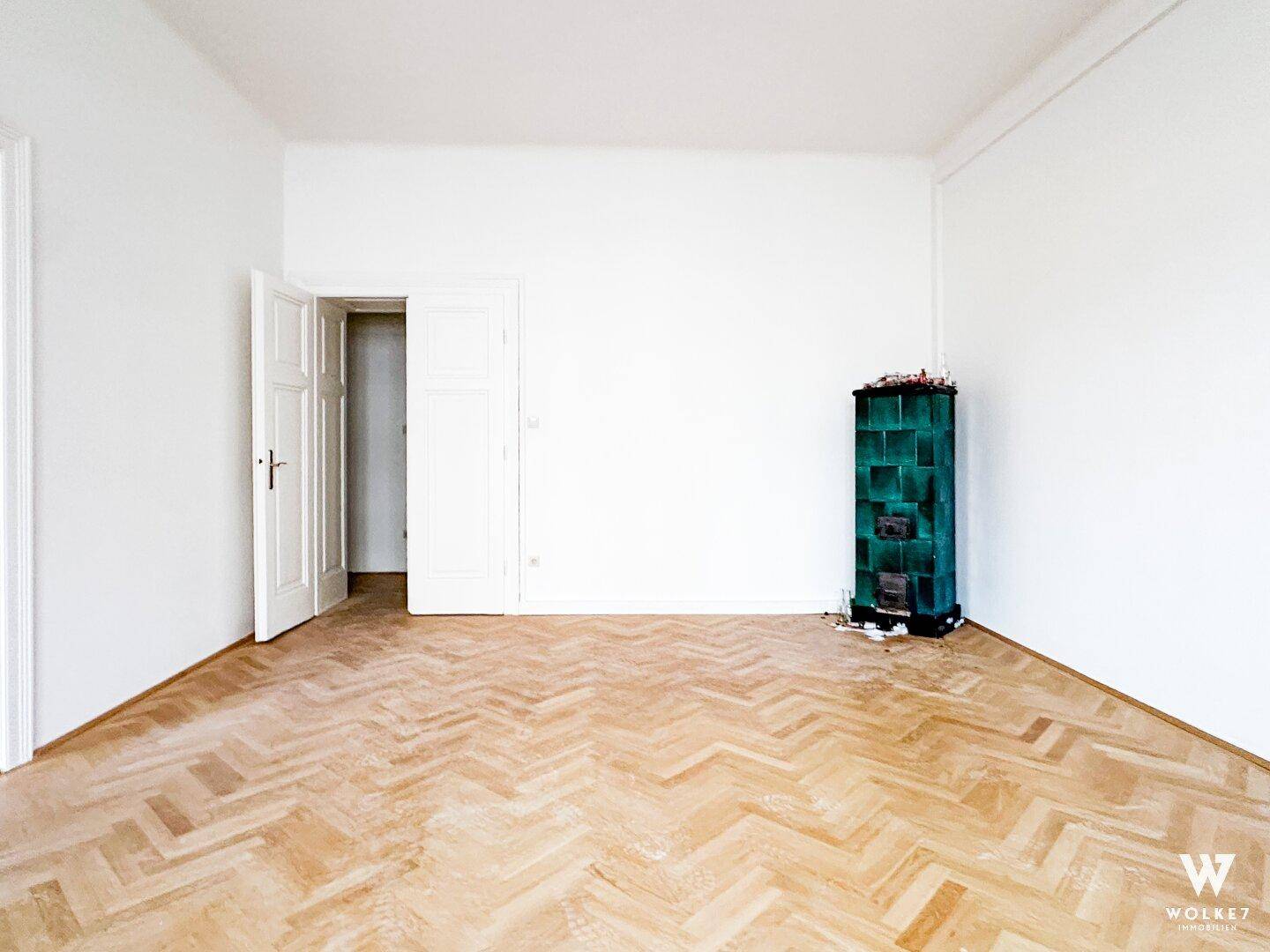 Geräumiger, leerer Raum mit Fischgrätparkett und einem traditionellen grünen Kachelofen an der Wand.