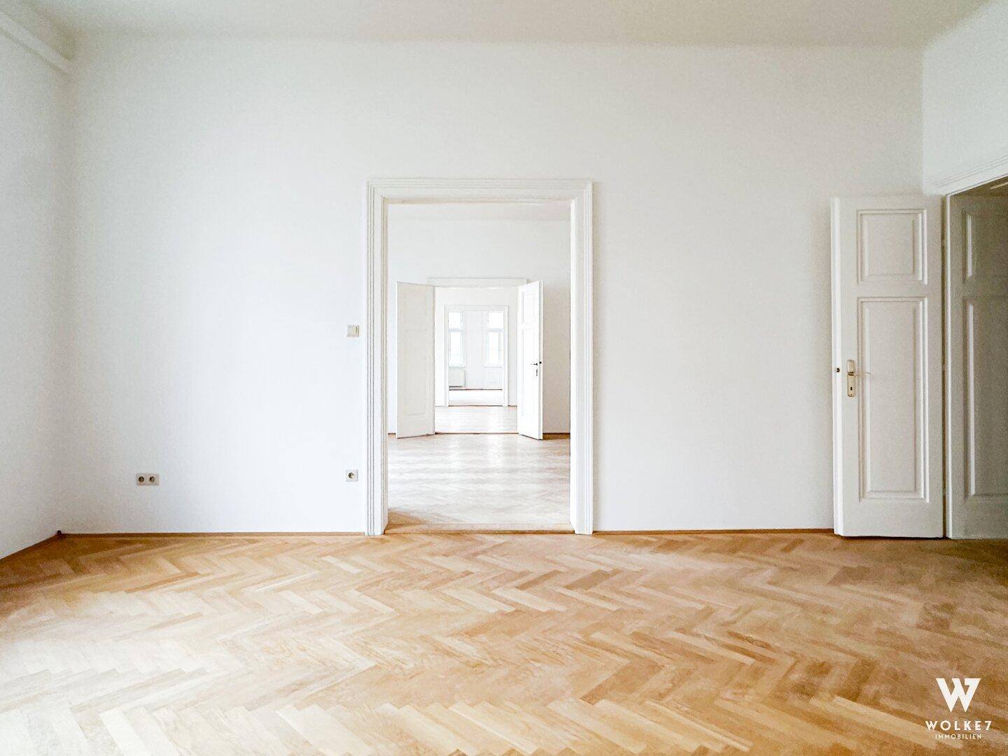 Heller, leerer Raum mit Fischgrätparkett und einer offenen Tür, die den Blick in einen weiteren Raum freigibt.