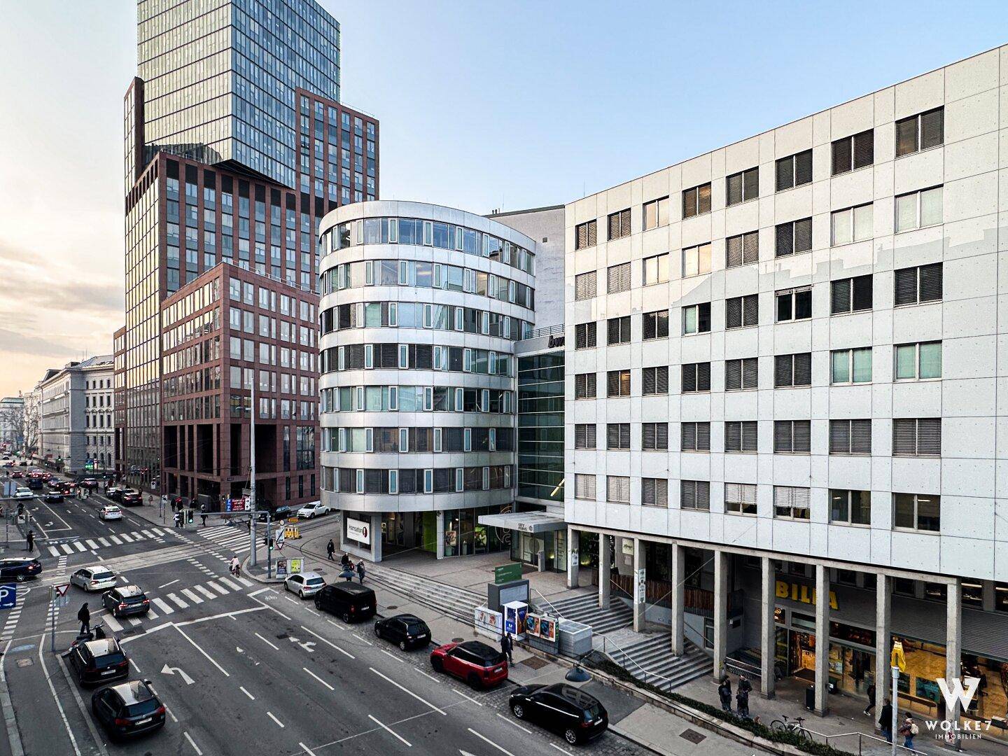 Moderne Stadtansicht mit verschiedenen Gebäuden, darunter ein Hochhaus und ein geschwungenes Gebäude, an einer belebten Kreuzung.