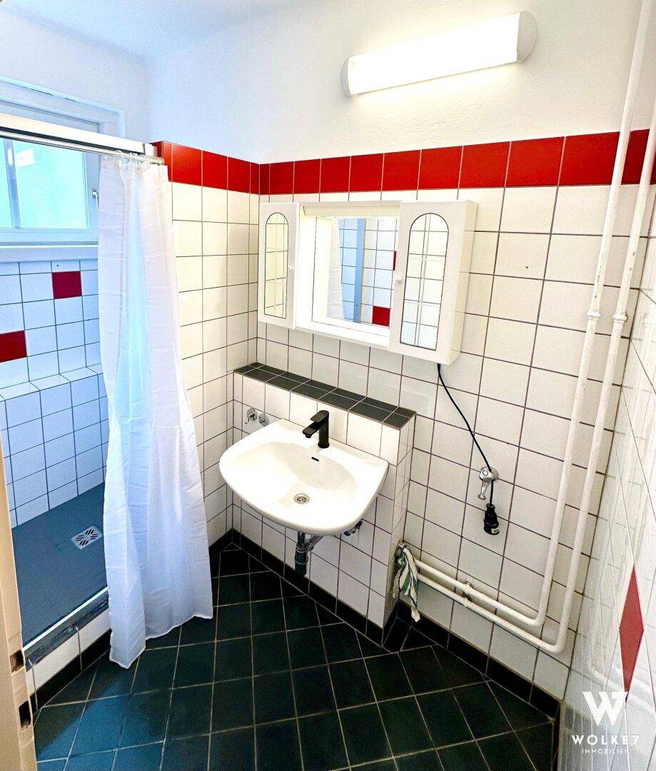 Badezimmer mit Dusche, Waschbecken und rot-weißen Fliesen, hell durch ein Fenster.