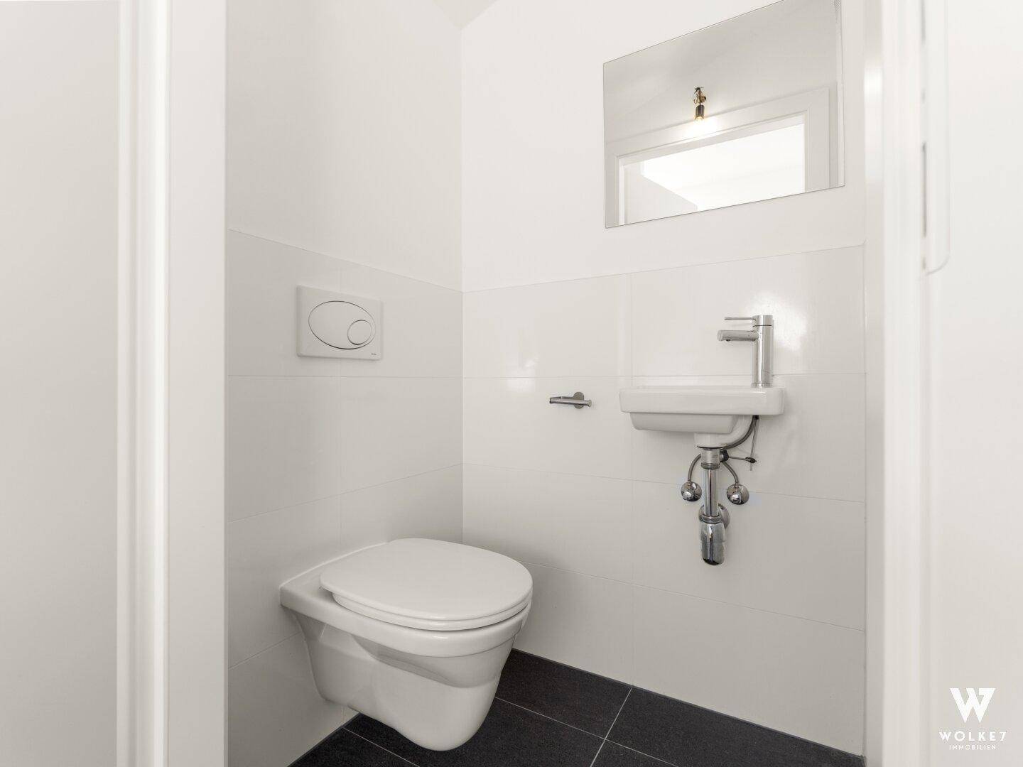Modernes Gäste-WC mit wandhängender Toilette, kleinem Waschbecken und dunklen Bodenfliesen.