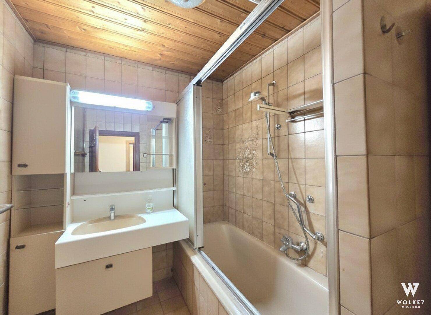 Älteres Badezimmer mit Badewanne, beigen Fliesen und Holzdeckenverkleidung, im Originalzustand.