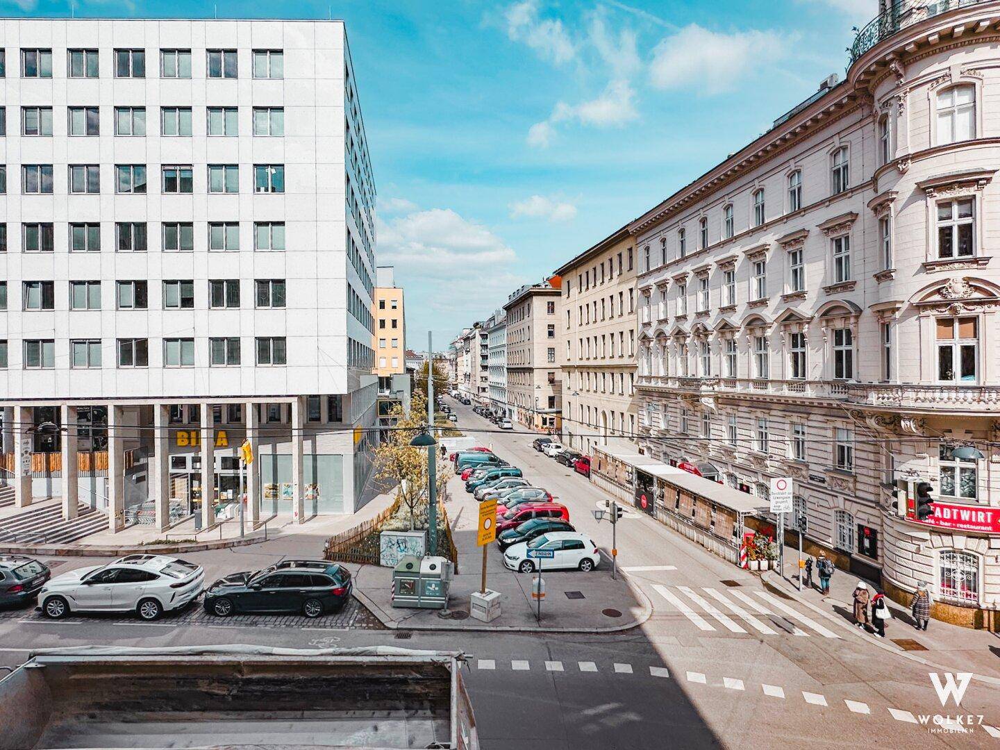 Stadtansicht mit Gebäuden, parkenden Autos und einer belebten Straße unter blauem Himmel.