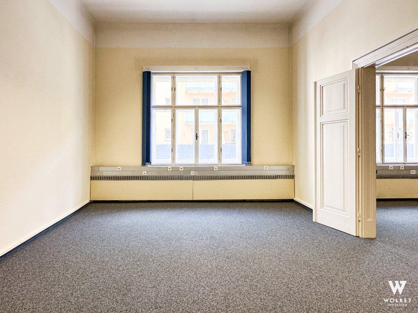 Leerer Raum in einer Arztpraxis mit Teppichboden und großen Fenstern, ideal für Behandlungszwecke.