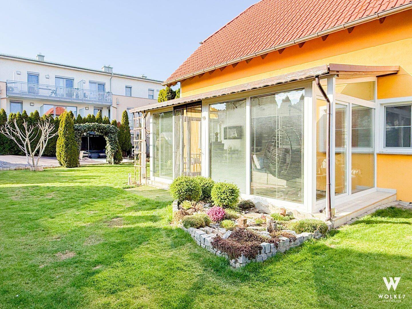 Gepflegter Gartenbereich mit Rasenfläche, Blumenbeet und einem Wintergarten am gelben Haus.