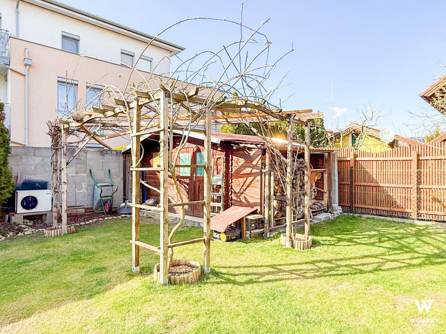 Sonniger Gartenbereich mit Holzschuppen, Rankgerüst und gepflegtem Rasen.