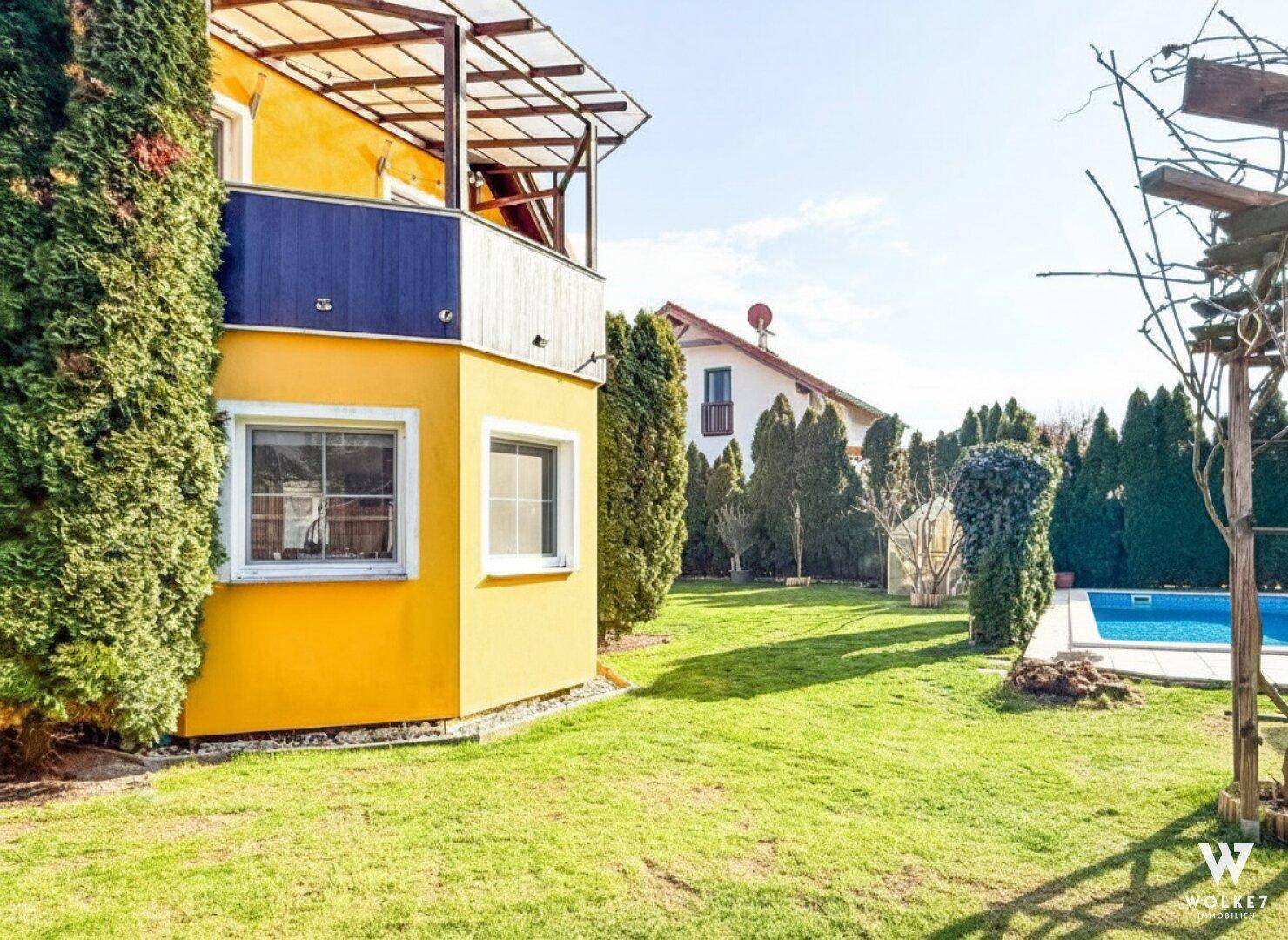 Gelbes Haus mit Balkon und Blick auf den gepflegten Garten mit Poolbereich.