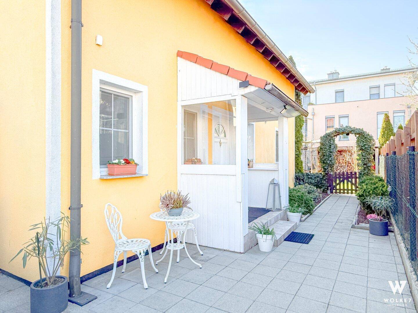 Eingangsbereich des Hauses mit überdachtem Vorbau, gepflastertem Weg und kleiner Sitzgelegenheit.