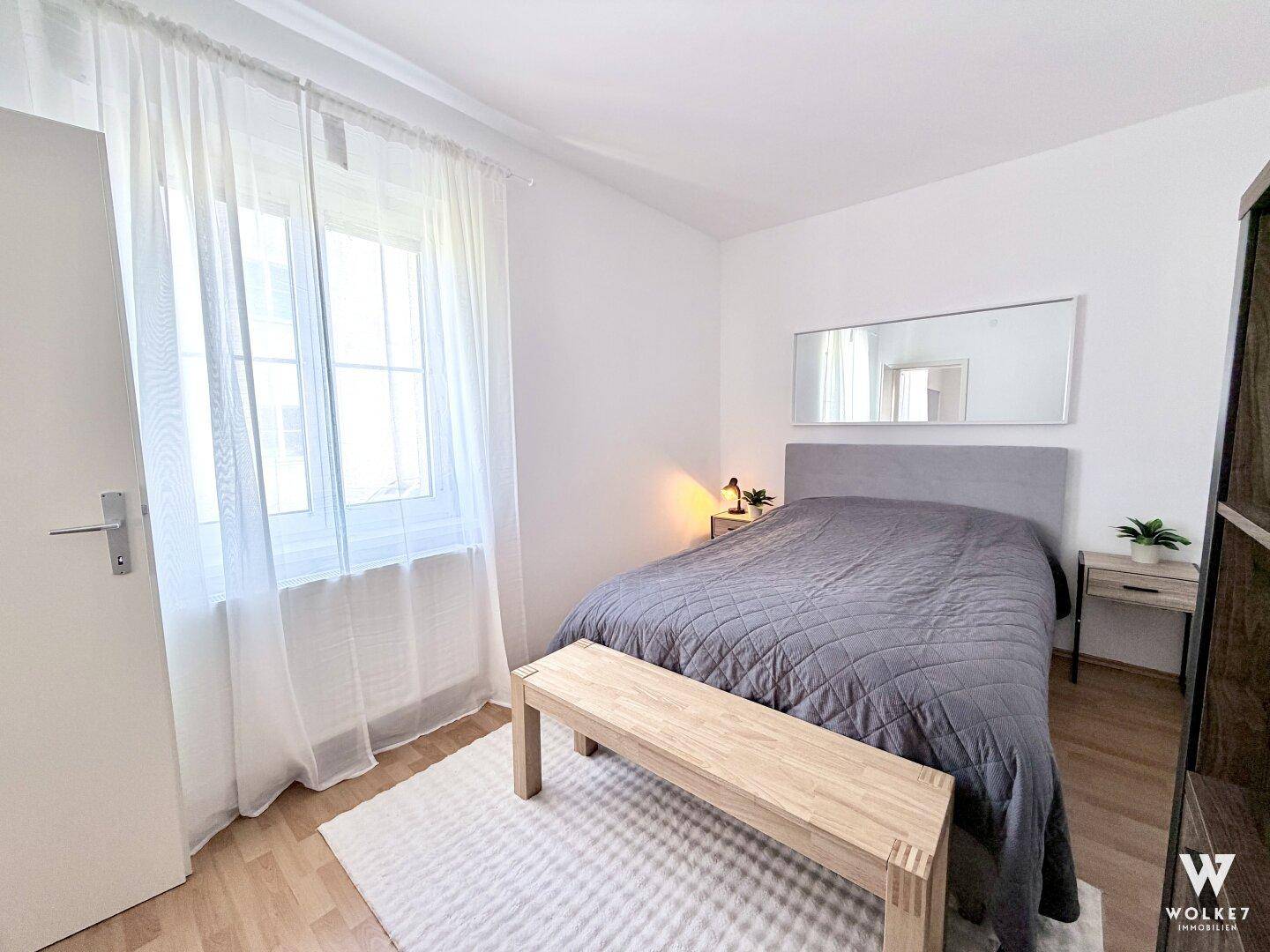 Schlafzimmer mit Doppelbett, Fenster und Holzbänken, hell und freundlich gestaltet.