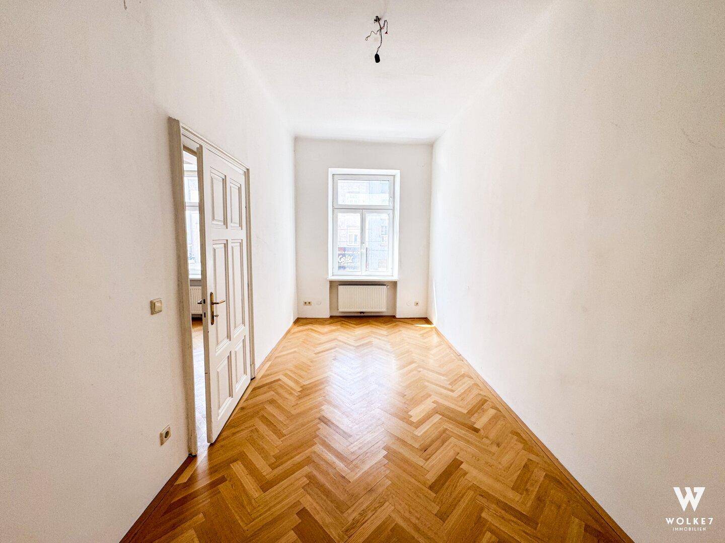 Schmaler, heller Raum mit Fischgrätparkett, einem Fenster und einer offenen Tür zu einem weiteren Zimmer.