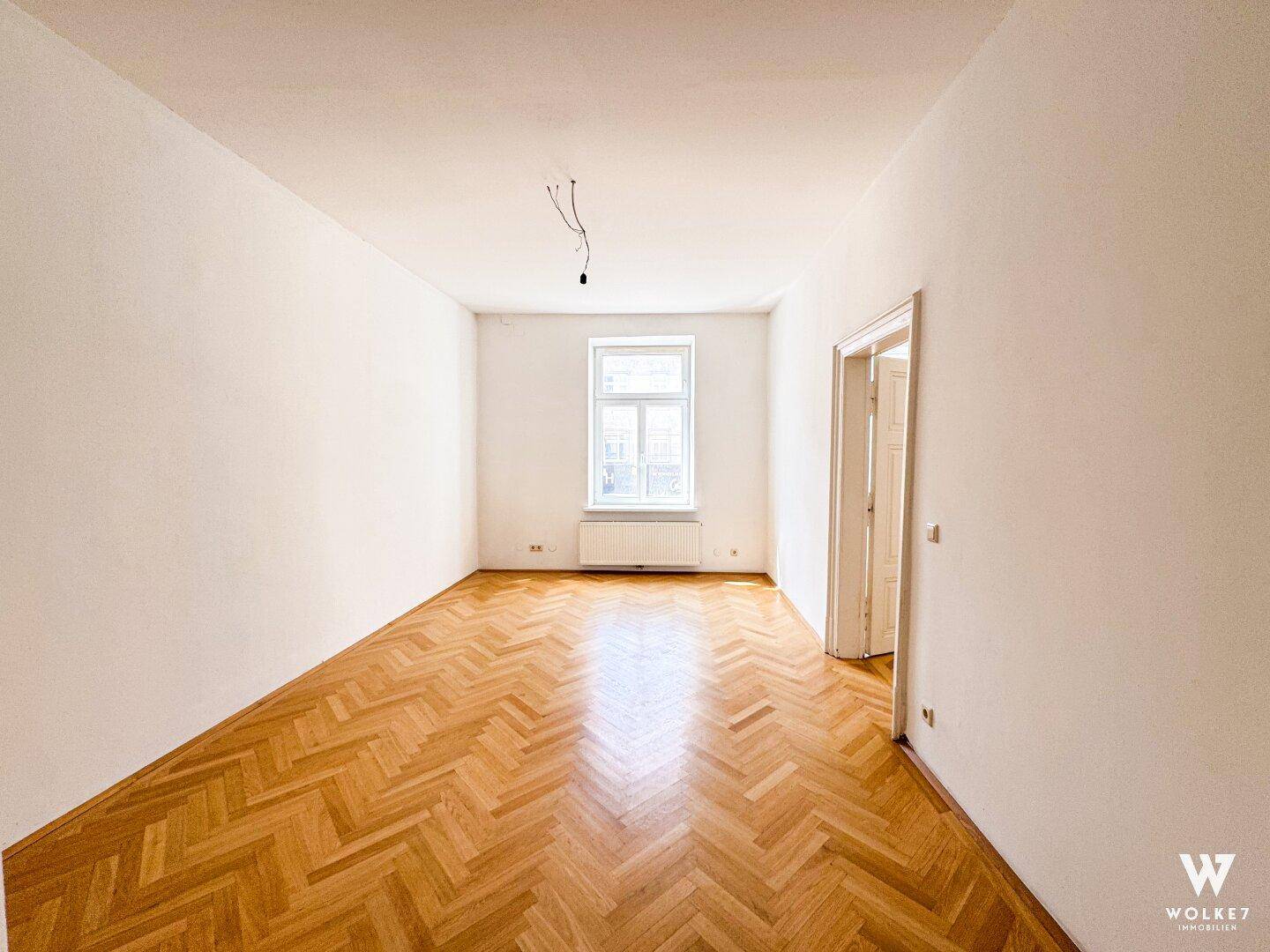 Heller, leerer Raum mit Fischgrätparkett, einem Fenster und einer offenen Tür, bereit zur individuellen Einrichtung.