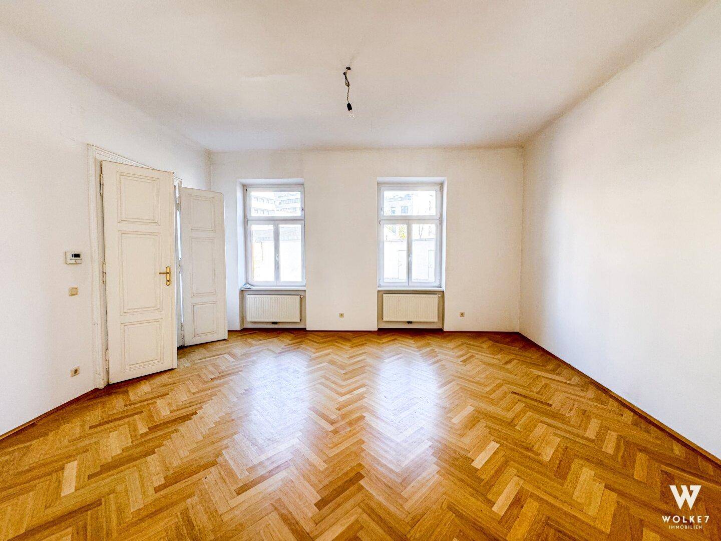 Geräumiger, leerer Raum mit Fischgrätparkett, zwei Fenstern und zwei offenen Türen, ideal für flexible Nutzung.