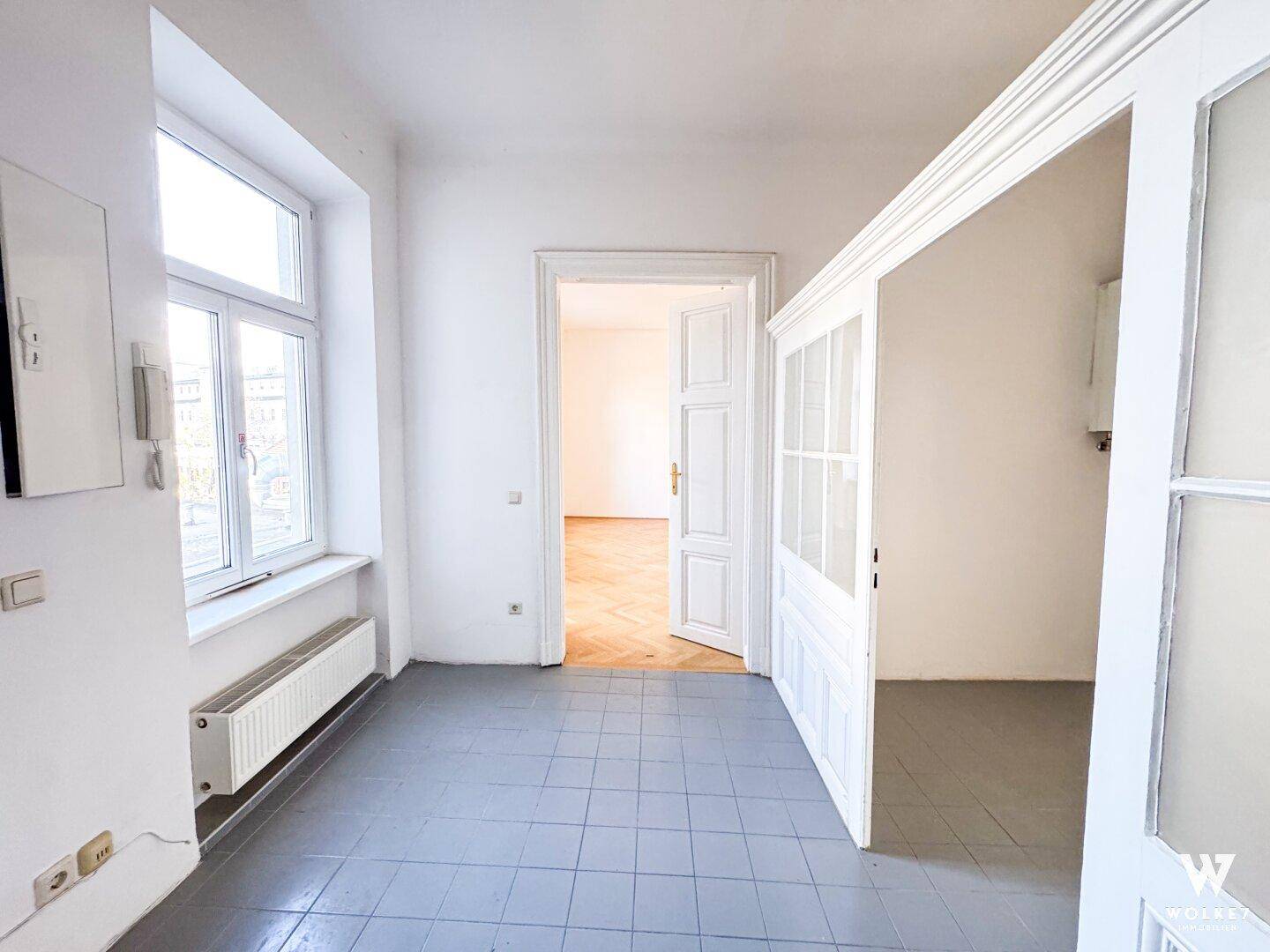 Eingangsbereich mit grauen Fliesen, einem Fenster und einer offenen Tür, die zu einem Raum mit Parkett führt.