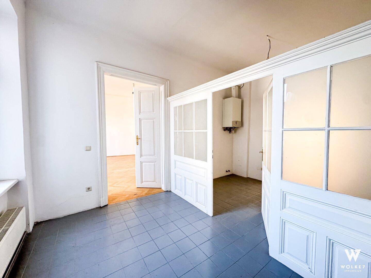 Eingangsbereich mit grauen Fliesen und einem Fenster, daneben ein offener Durchgang zu einem weiteren Zimmer.