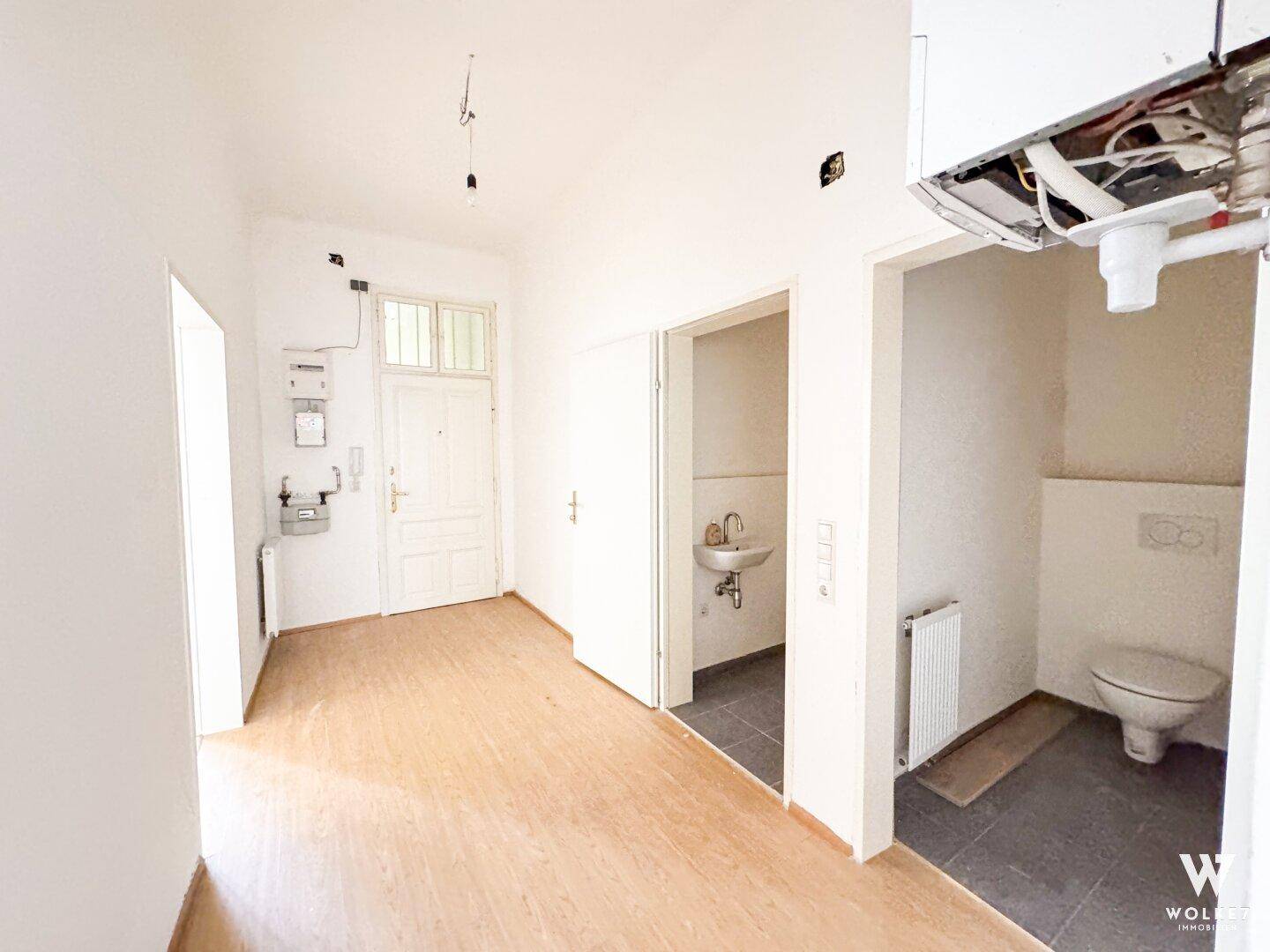 Eingangsbereich mit Zugang zu Toilette und weiteren Räumen, heller Laminatboden und weiße Wände.