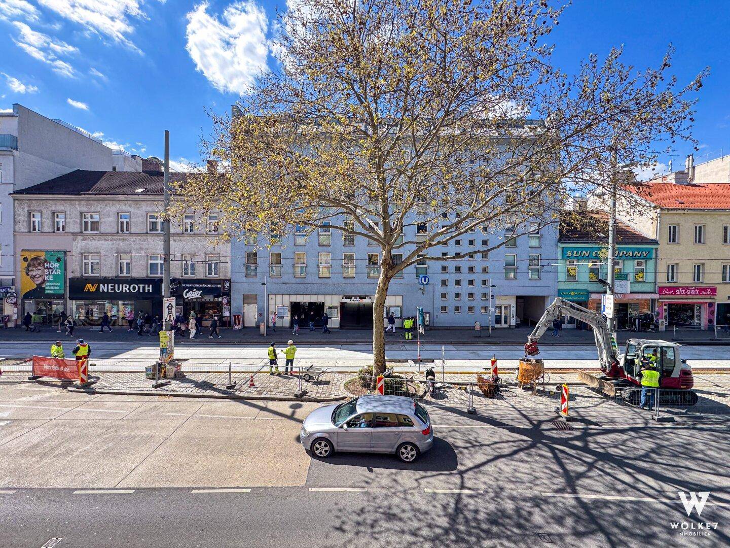 Stadtansicht mit Gebäuden, Bäumen und Bauarbeiten auf der Straße unter blauem Himmel.