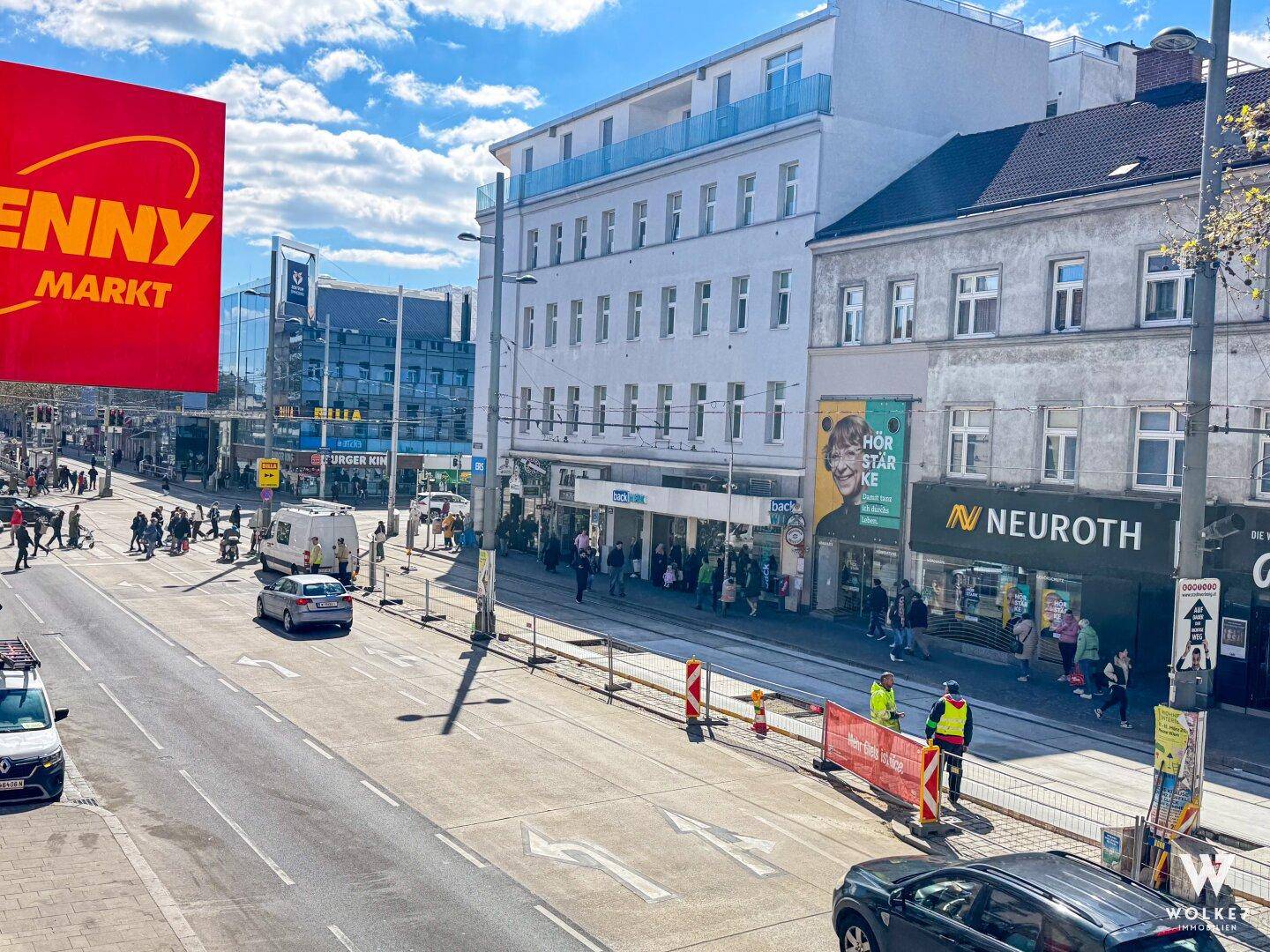 Weitläufige Straßenansicht mit regem Verkehr, Geschäften und Passanten, die das städtische Leben widerspiegelt.
