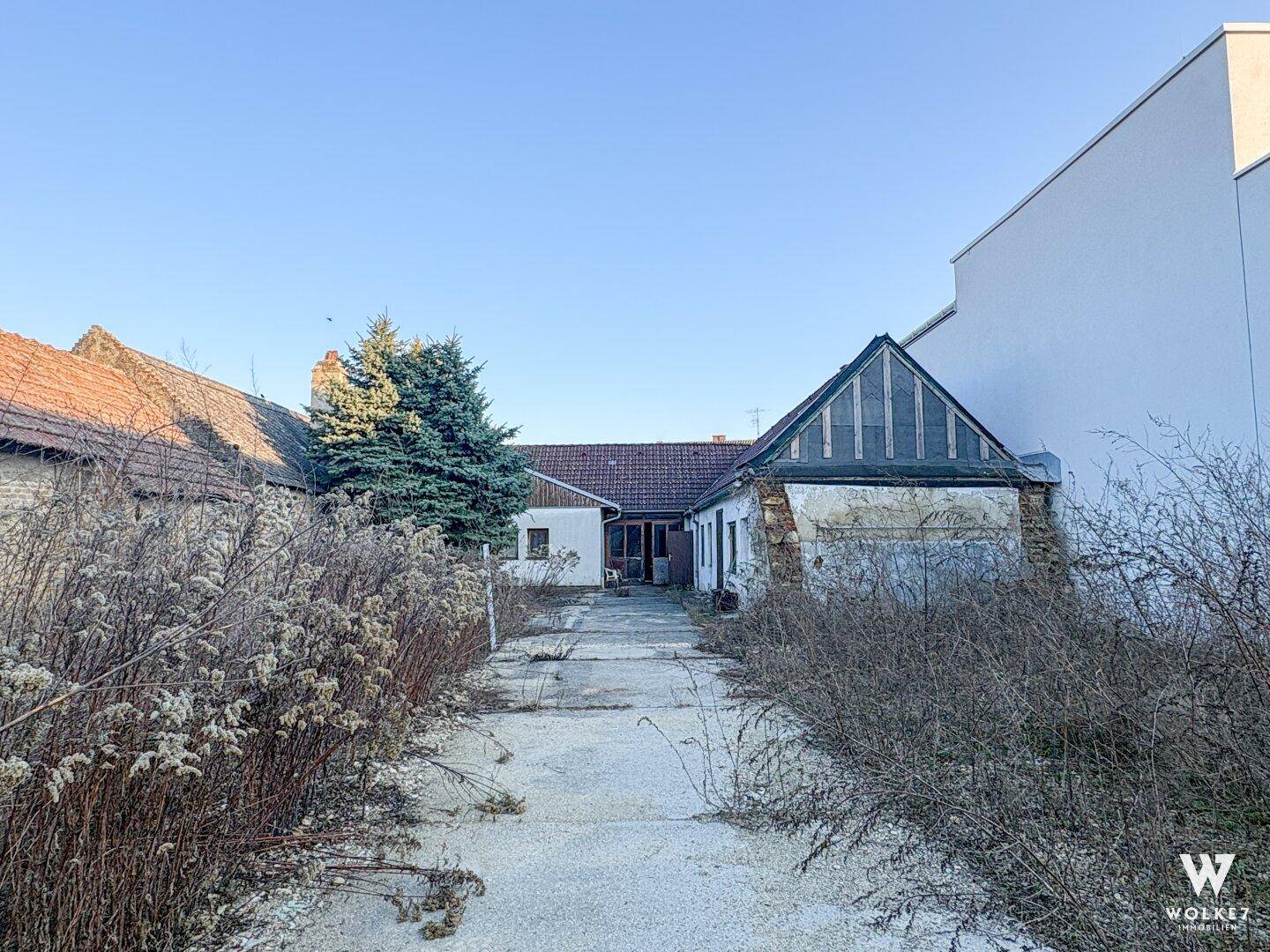 Langer Weg zwischen Gebäuden mit viel Wildwuchs und Blick auf weitere Gebäude im Hintergrund.