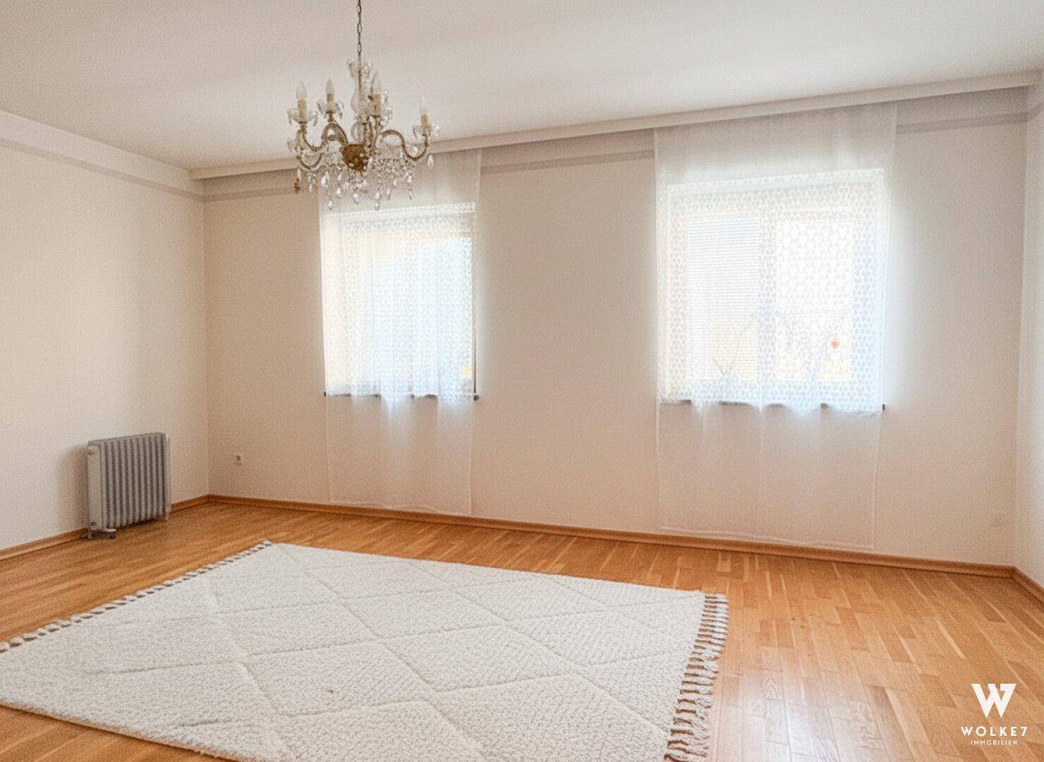 Helles Zimmer mit Parkettboden, zwei Fenstern und einem eleganten Kronleuchter an der Decke.