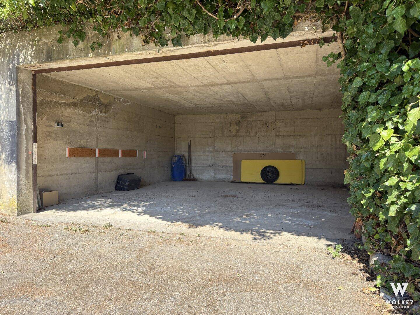 Offene Garage mit Betonboden, umgeben von Grünflächen und Efeu.