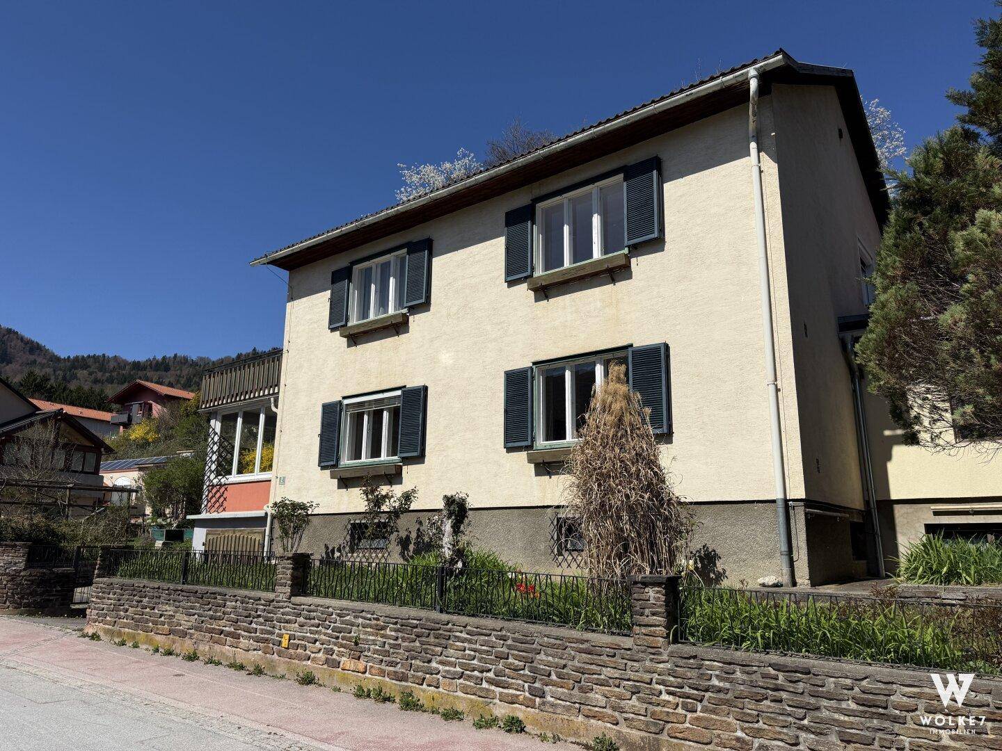 Seitenansicht des Wohnhauses mit grünen Fensterläden und Steinmauer im Vorgartenbereich.