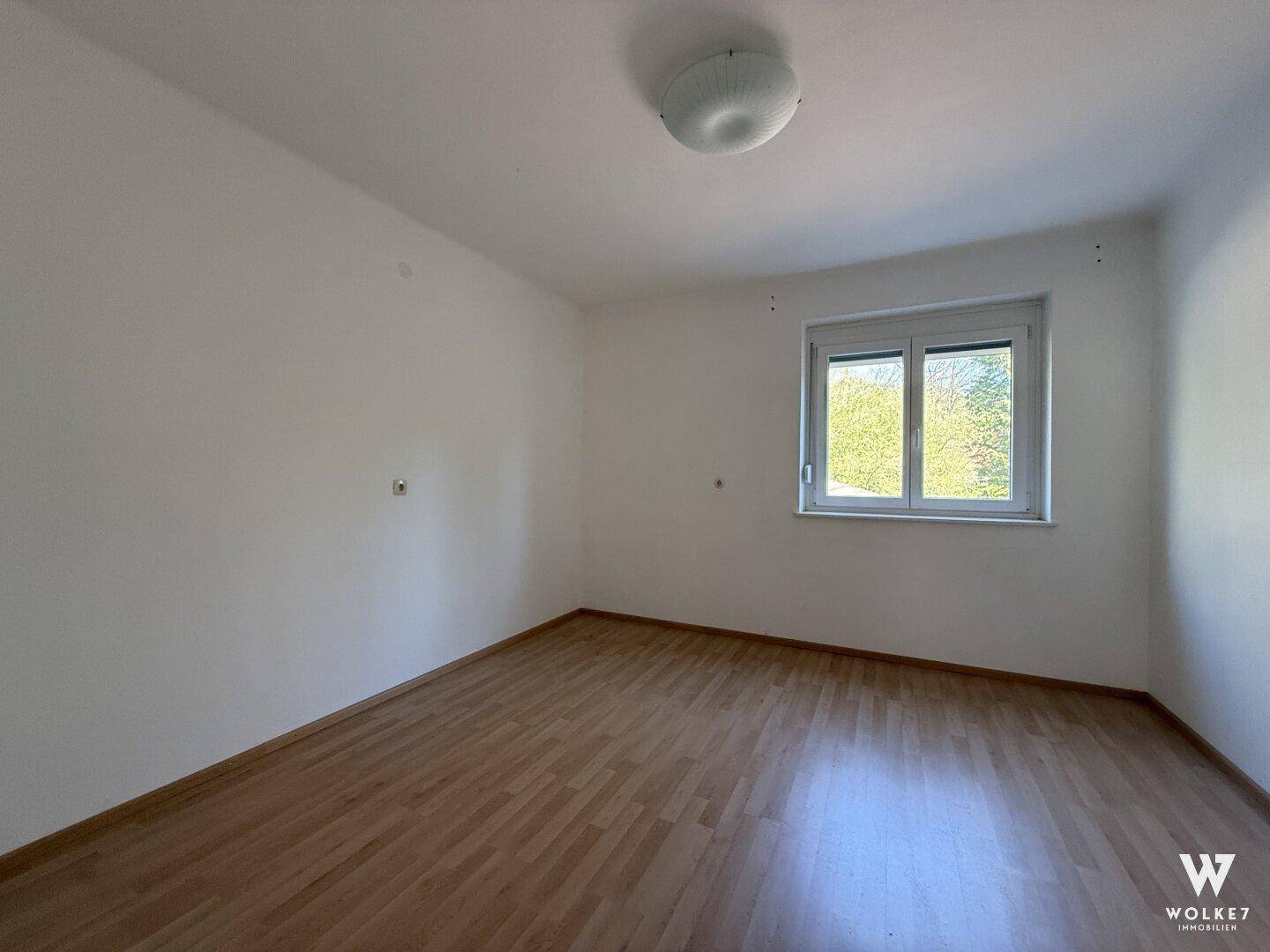 Einfaches Schlafzimmer mit Laminatboden und Fenster mit Blick ins Grüne.