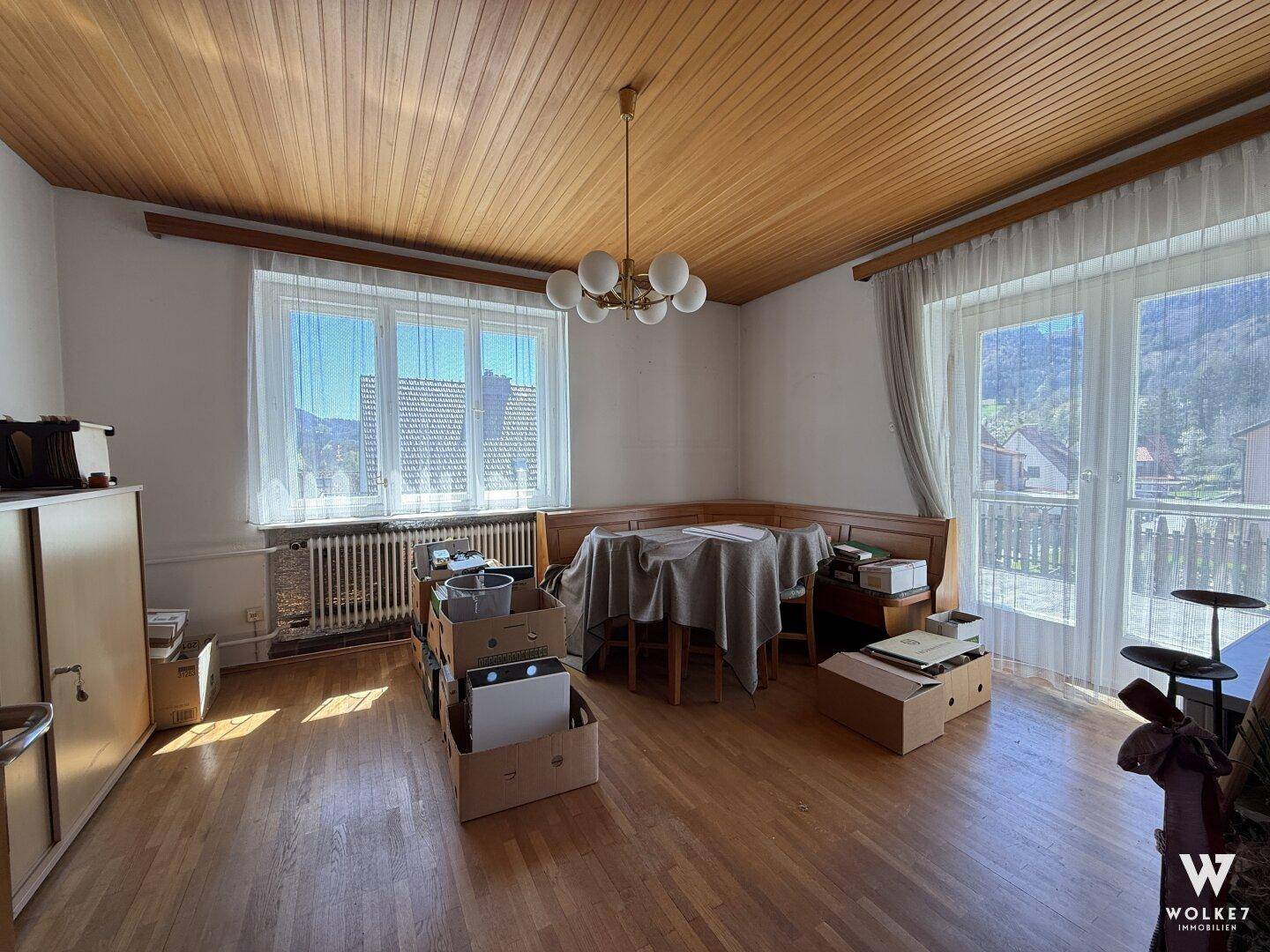 Essbereich mit Holzdecke, großem Fenster und Zugang zu einem sonnigen Balkon.