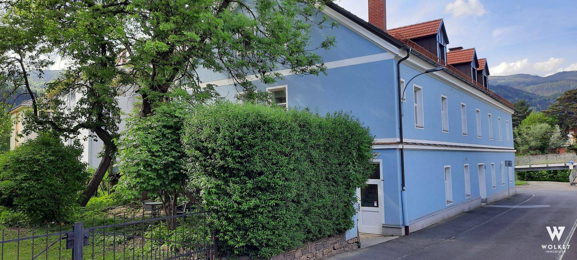 Seitenansicht des blauen Gebäudes mit rotem Dach, umgeben von Hecken und Bäumen, mit Bergen im Hintergrund.