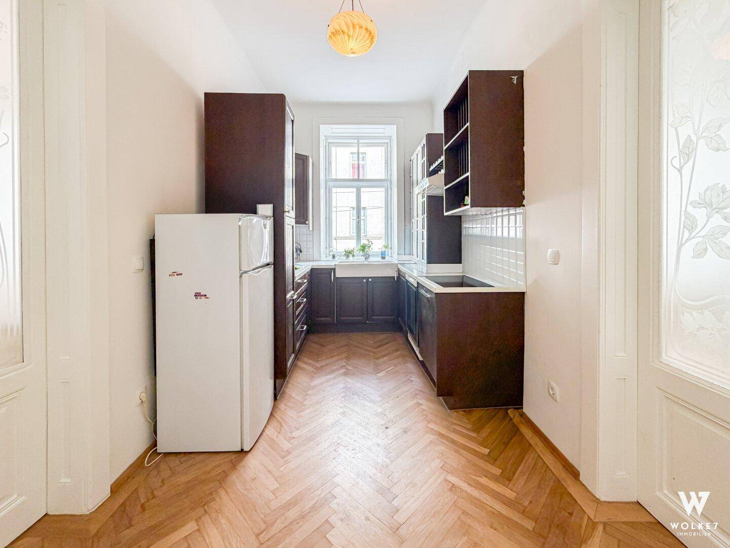 Schmale Küche mit dunklen Holzschränken, Kühlschrank und Fischgrätparkettboden.