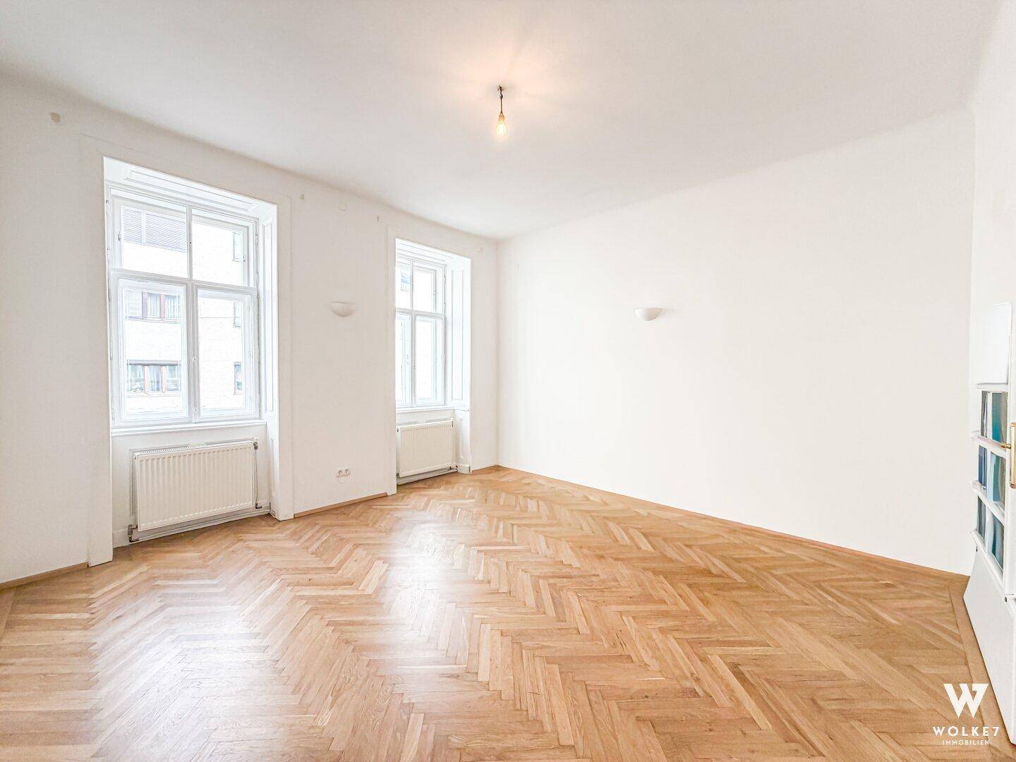 Leeres Wohnzimmer mit hellem Fischgrätparkett und großen Fenstern in einem Altbau.