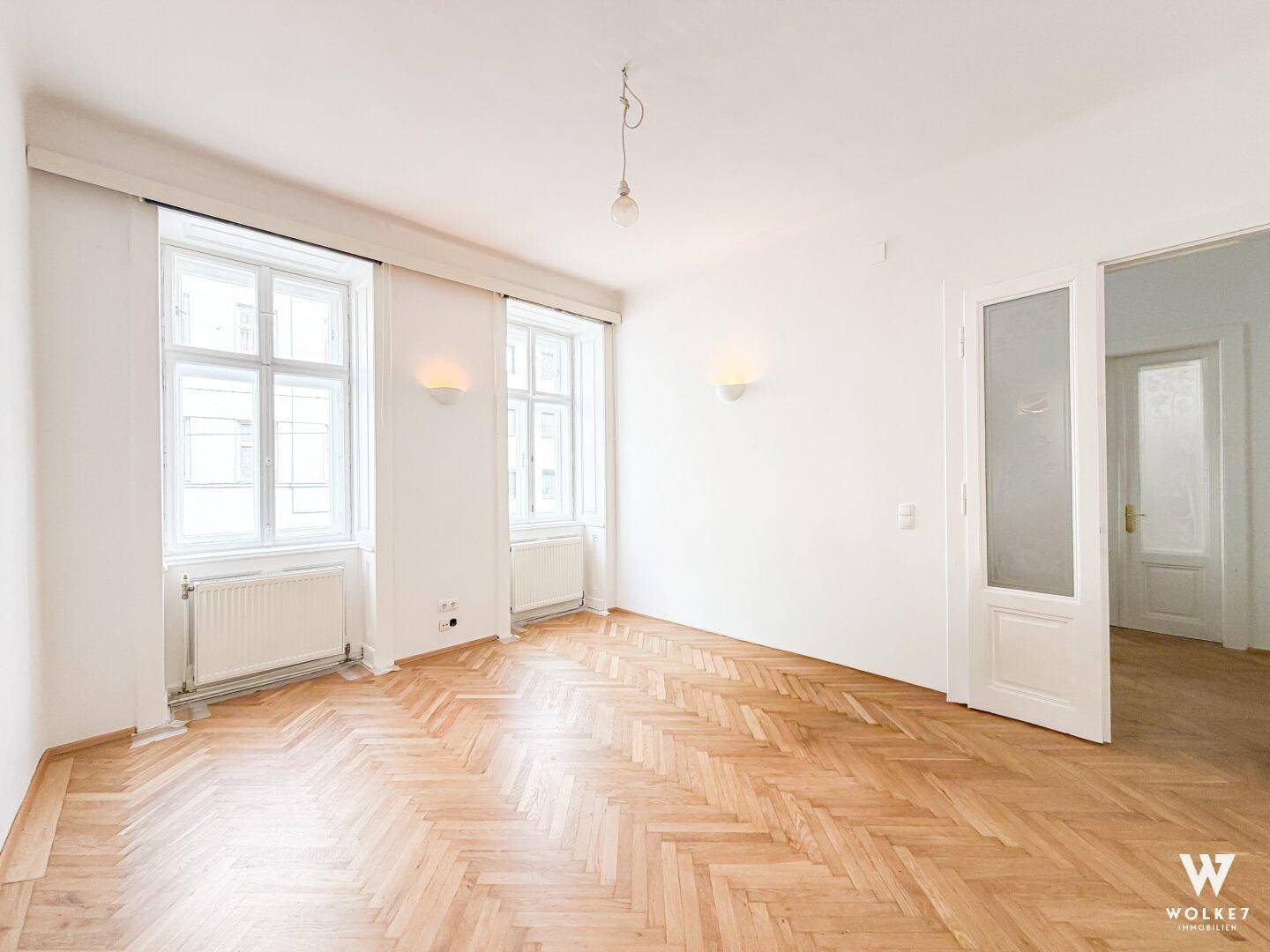 Geräumiges, leeres Wohnzimmer mit Fischgrätparkett, hohen Fenstern und weißen Wänden.