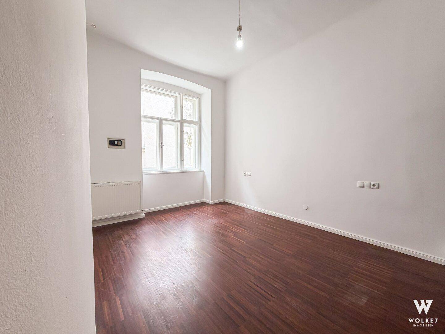 Leeres Schlafzimmer mit dunklem Holzboden und großem Fenster in einem Altbau.